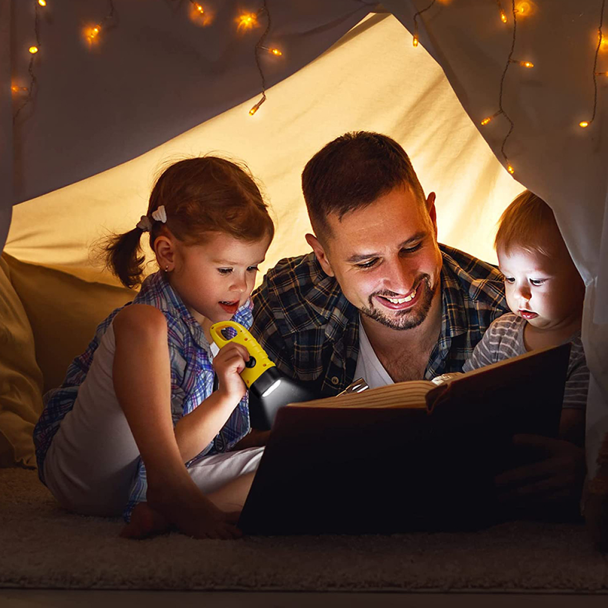 Eine Familie liest unter einem Zelt mit Lichterketten ein Buch. Ein Kind hält eine Taschenlampe.