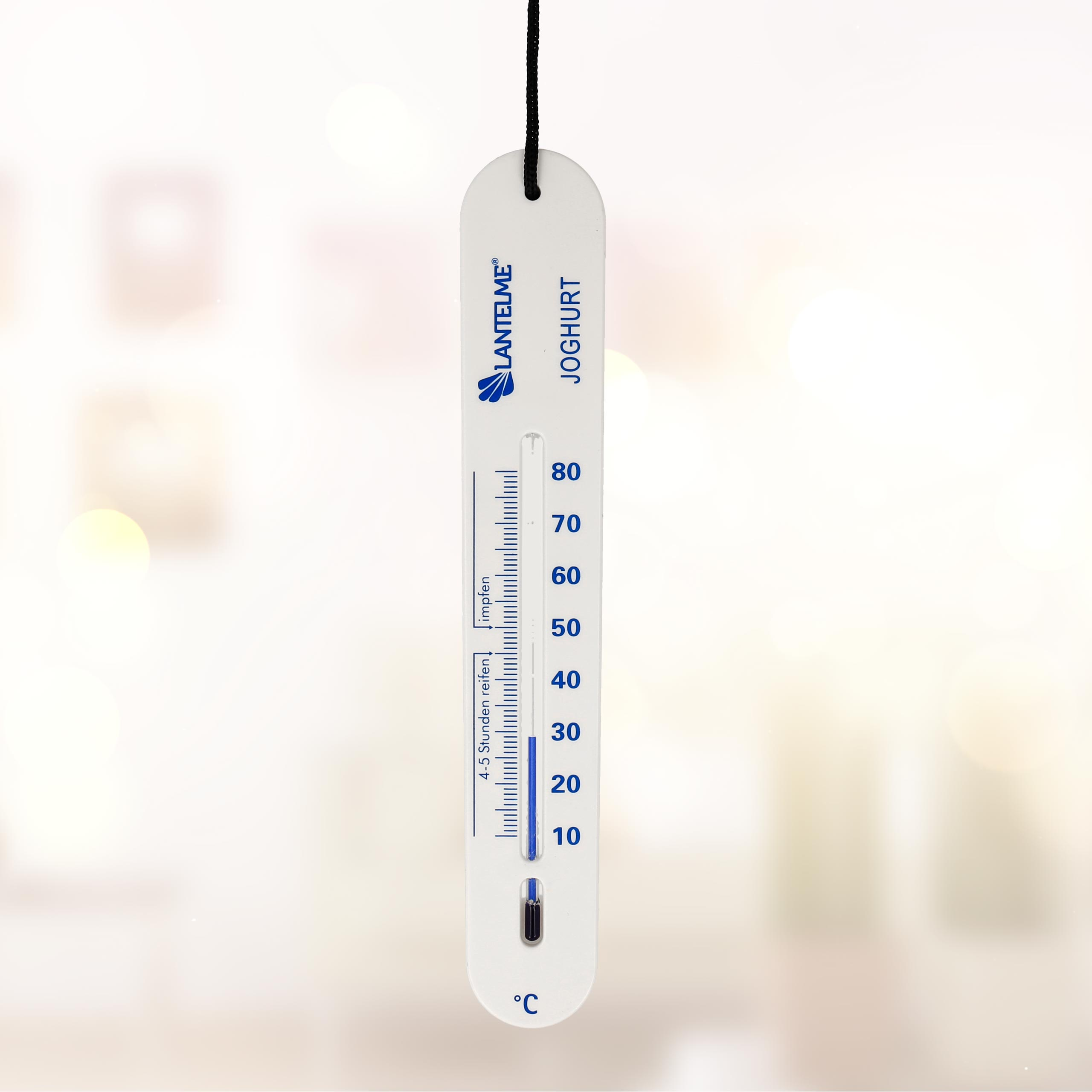 Ein weißes Thermometer mit blauen Markierungen hängt an einer Schnur. Der Hintergrund ist unscharf.