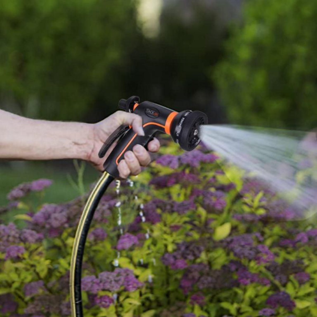 Eine Hand hält eine Wasserpistole und sprüht Wasser auf lila Blumen. Der Hintergrund ist grünes Laub.