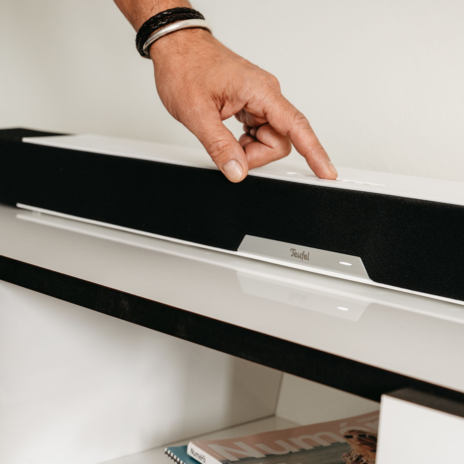 Eine Hand drückt eine Taste auf einer weißen und schwarzen Soundbar mit dem Teufel-Logo auf der Vorderseite.