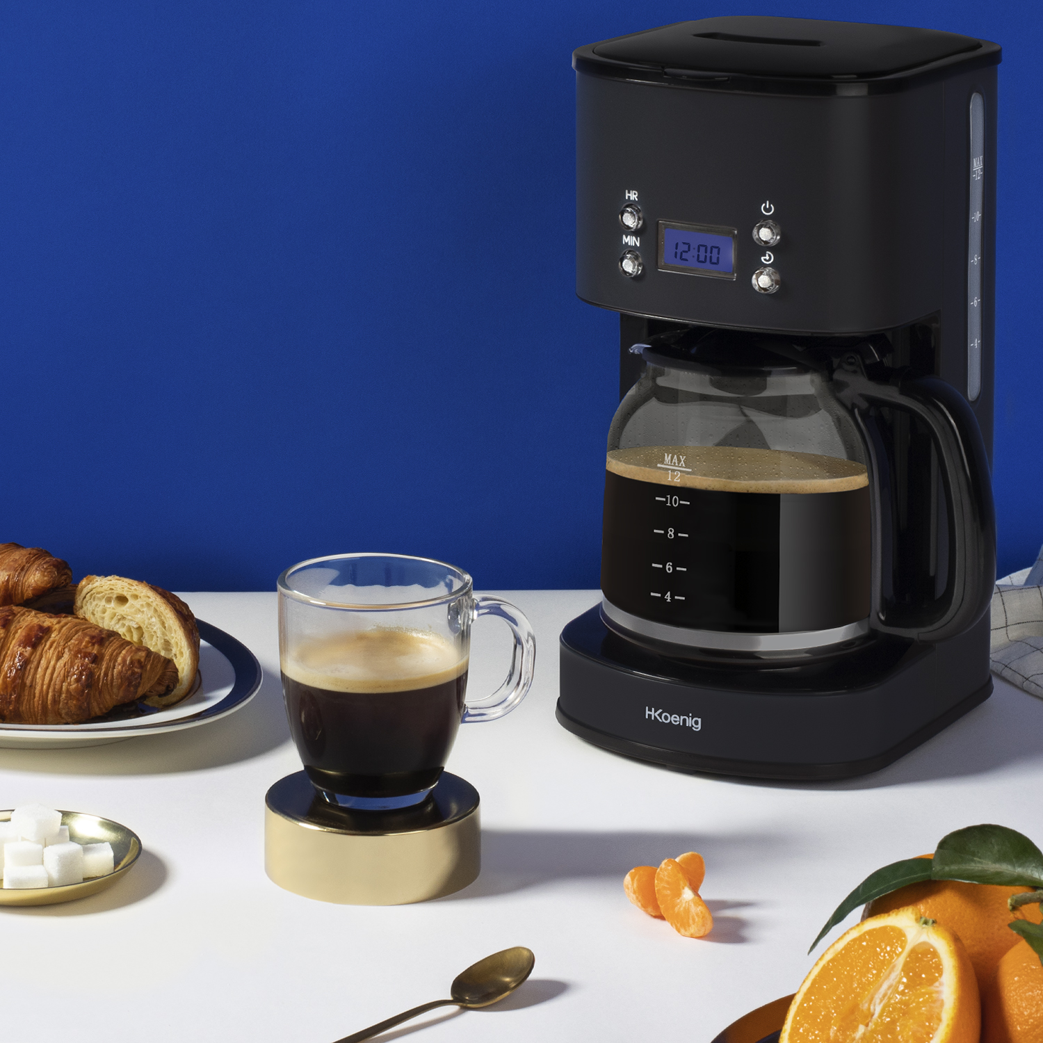Zwarte koffiezetapparaat met een glazen kan en een kopje koffie. Er zijn ook croissants en sinaasappels.
