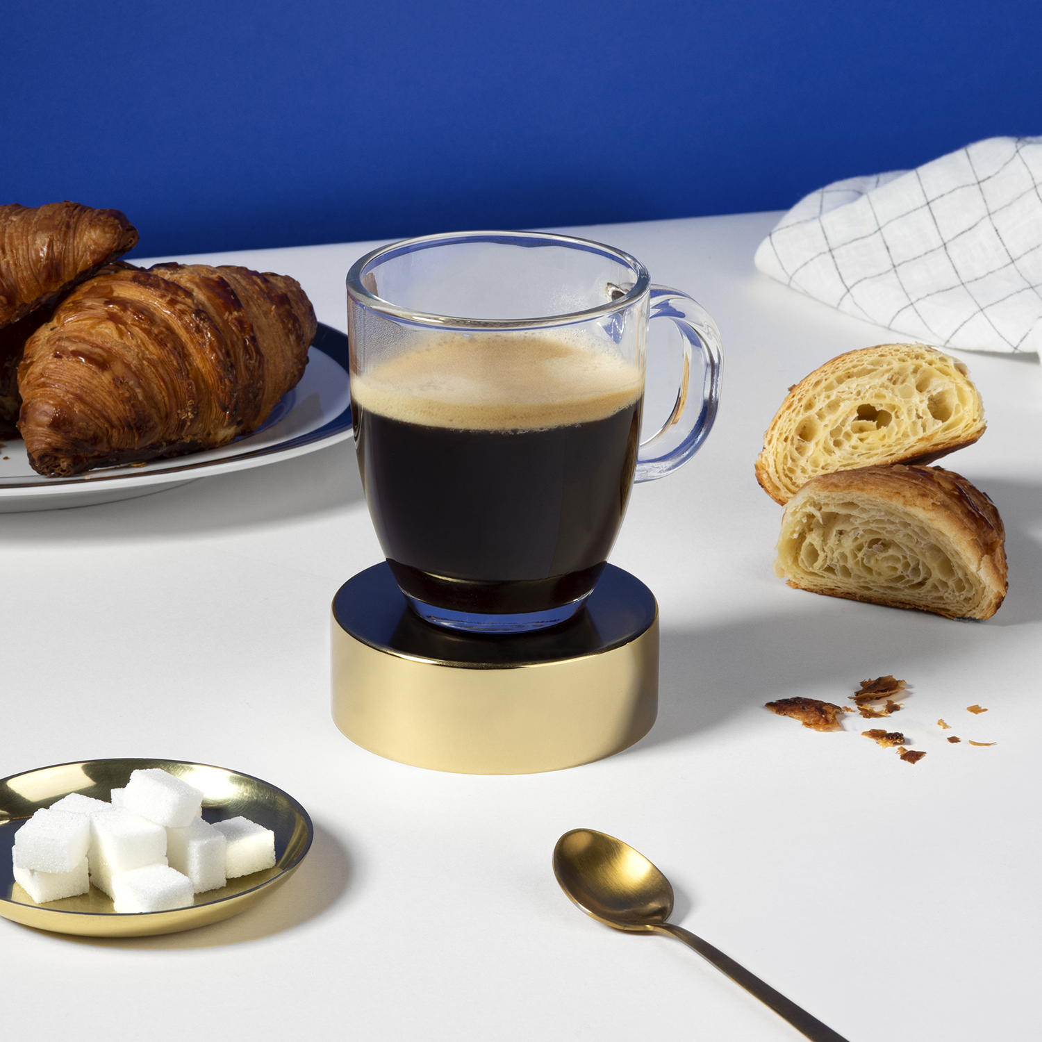 Koffie in een glazen mok op een standaard, croissants, suikerklontjes en een lepel.