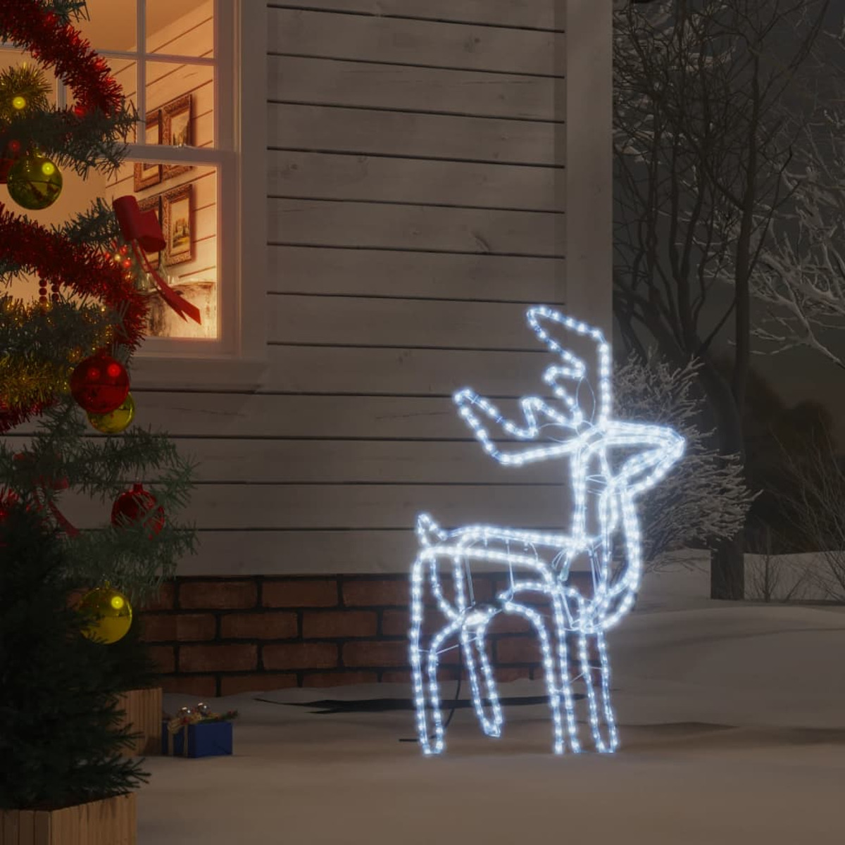 Eine leuchtende weiße Rentierdekoration steht auf verschneitem Boden vor einem Haus. Weihnachtsbeleuchtung und Baum sichtbar.