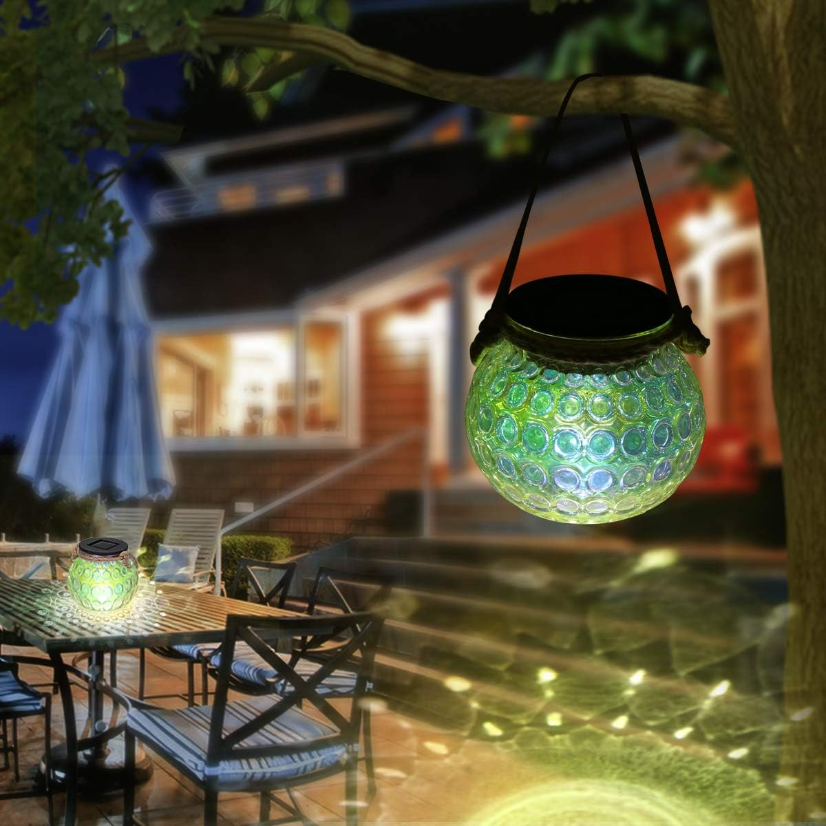 Eine leuchtende Glaskugel hängt an einem Ast. Die Kugel und eine andere auf einem Tisch beleuchten eine Terrasse mit einem Haus im Hintergrund.