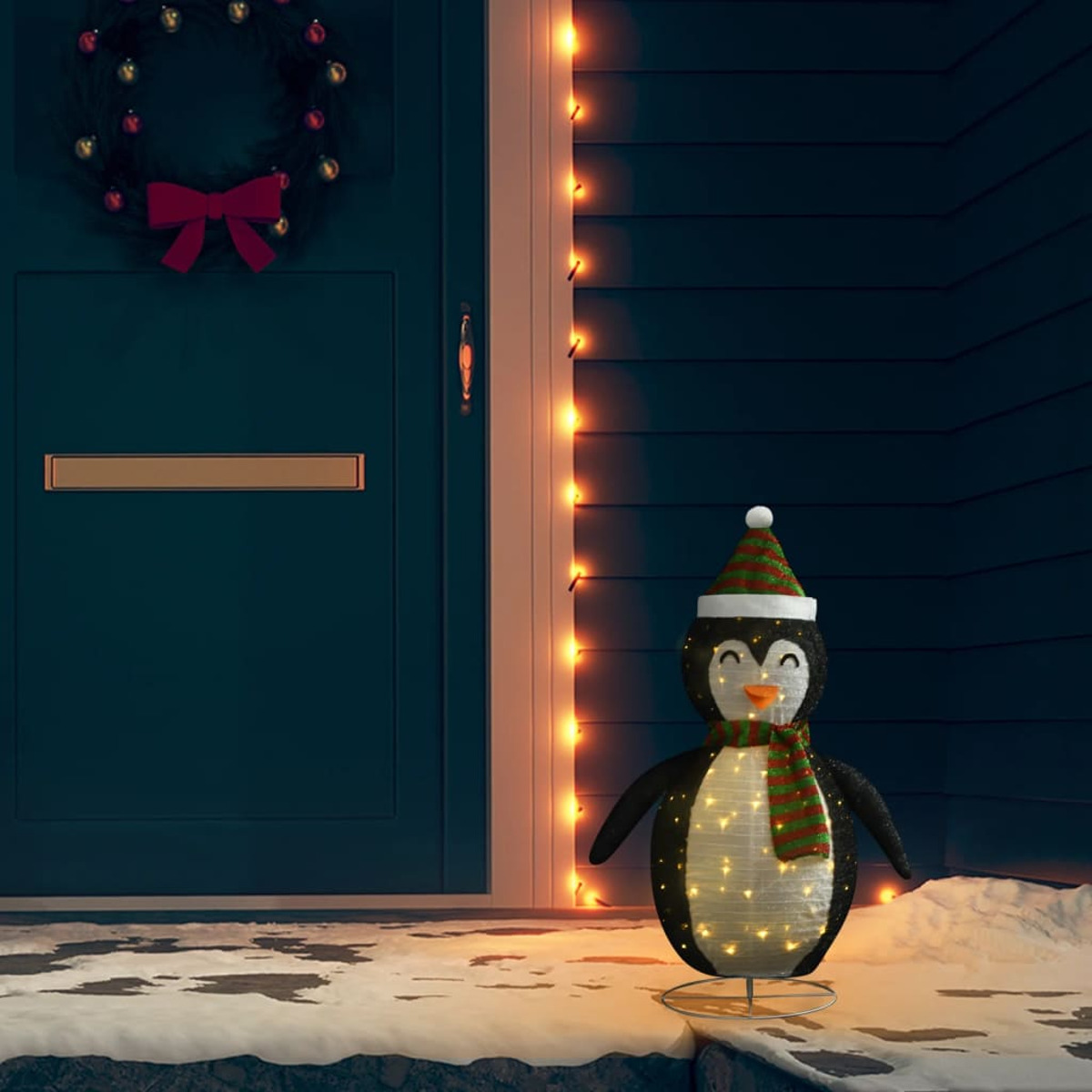 Weihnachtsdekoration Pinguin mit Lichtern. Steht im Schnee, vor Tür mit Kranz.