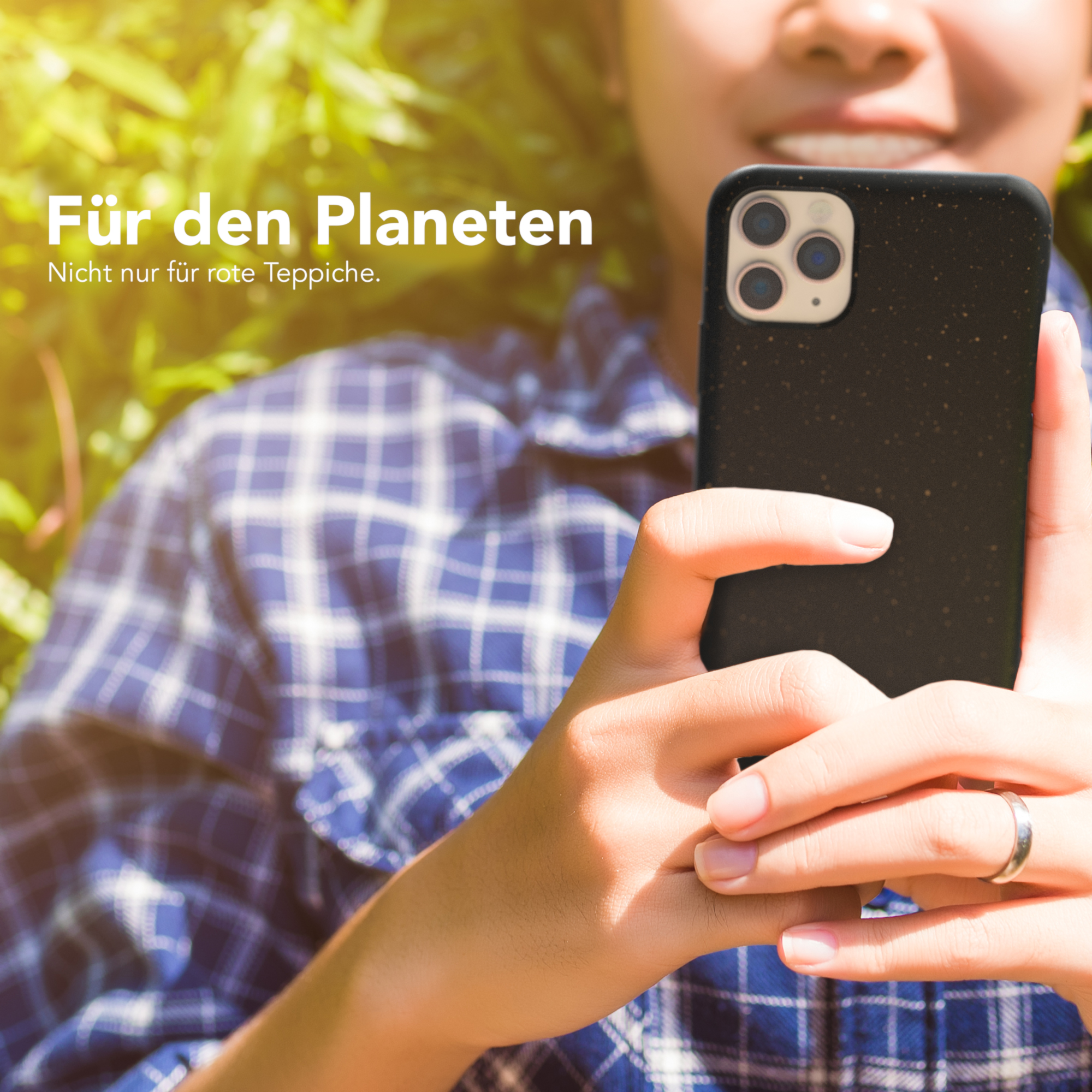 Person hält ein schwarzes Telefon mit Hülle, grüne Blätter im Hintergrund.