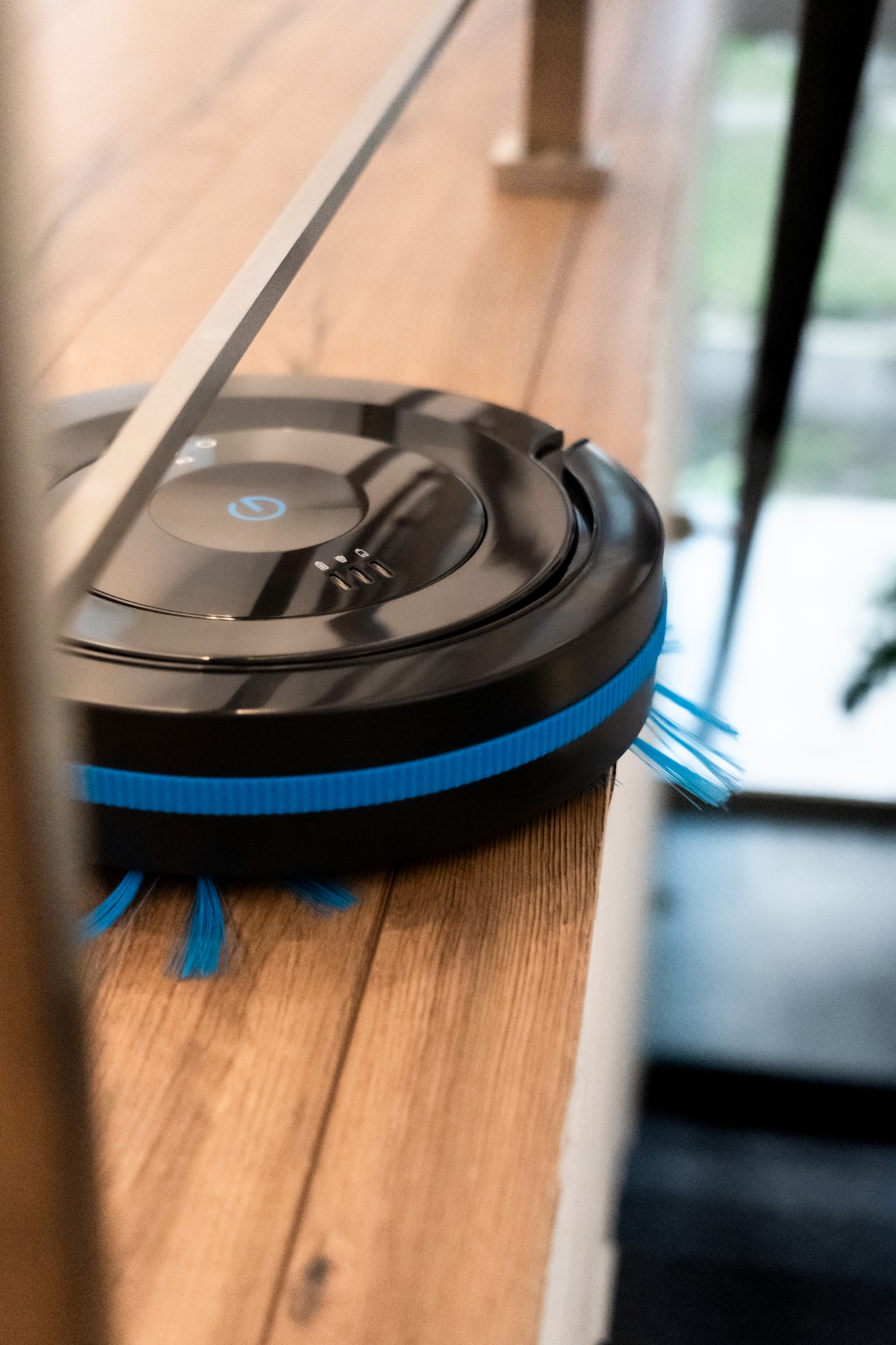 Ein schwarz-blauer Roboter-Staubsauger auf einem Holzboden, in der Nähe einer Holzkante.