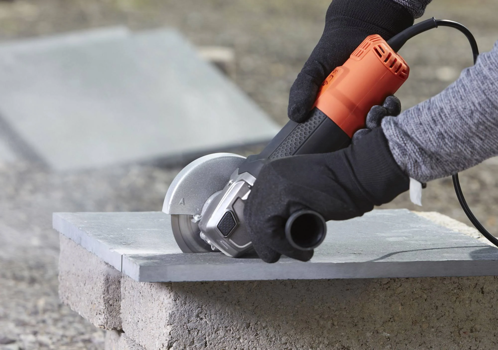 Dłonie w czarnych rękawicach używają szlifierki na szarym kamieniu. Tło jest rozmyte.