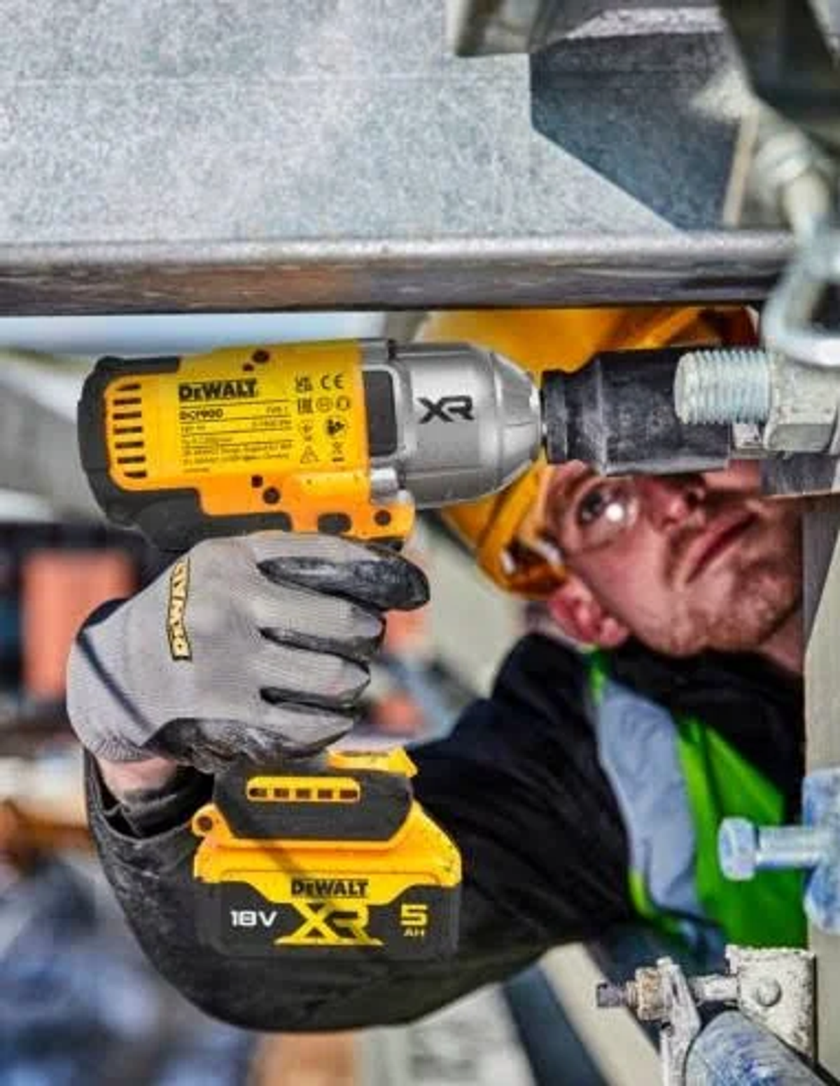 Robotnik używający żółto-czarnej wiertarki na metalu, w kasku, okularach ochronnych i rękawicach.