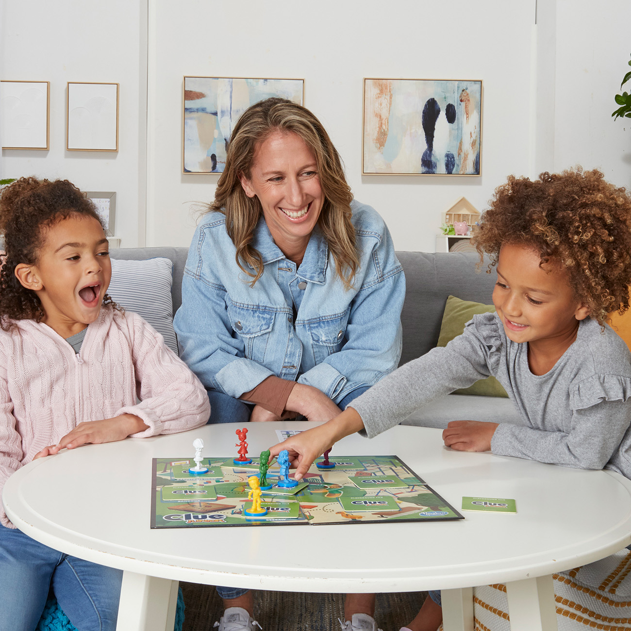 Eine Familie spielt Clue. Eine Mutter beobachtet zwei aufgeregte Mädchen an einem runden Tisch.
