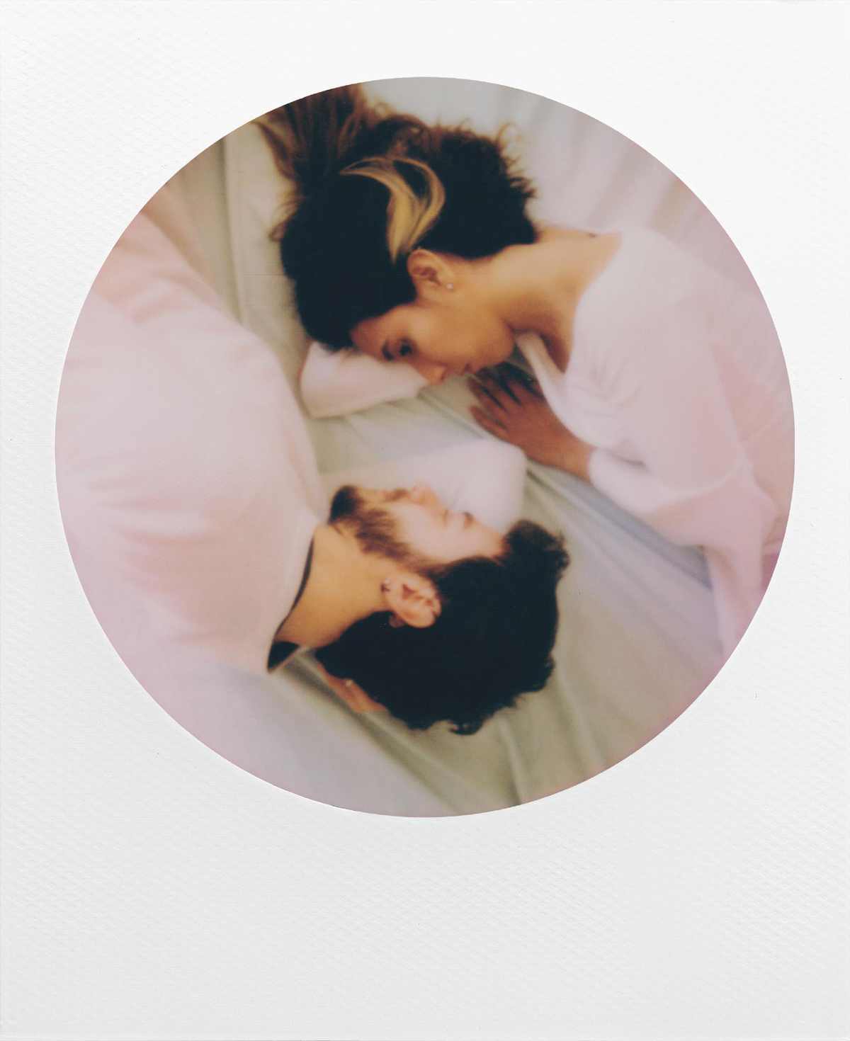 Ein Polaroid-Bild von einem Mann und einer Frau, die Kopf an Kopf auf einem hellblauen Bett liegen. Das Bild ist in einem runden Rahmen.