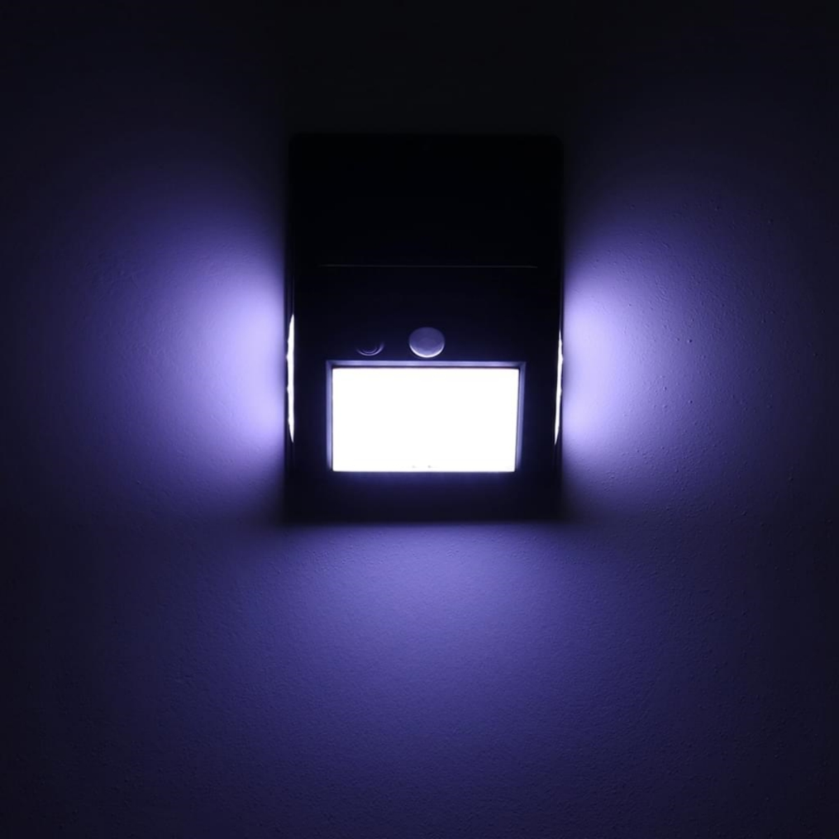 Eine Wandleuchte mit rechteckiger Form strahlt ein helles, weißes Licht aus, umgeben von einem violetten Schein, in einem dunklen Raum.