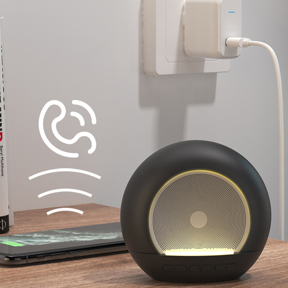 Ein schwarzer, runder Lautsprecher neben einem ladenden Telefon. Ein Wandstecker ist im Hintergrund. Schallwellen-Symbol.