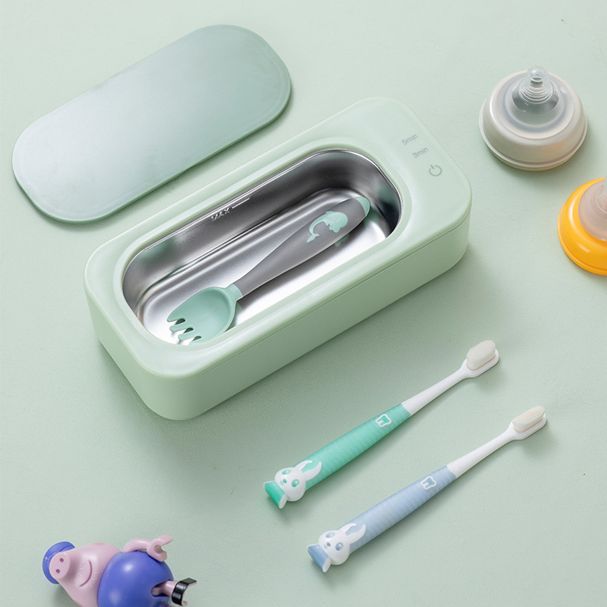 Ein Babyflaschensterilisator mit Löffel und Gabel, zwei Zahnbürsten und Babyflaschenzubehör liegen auf einer Oberfläche.