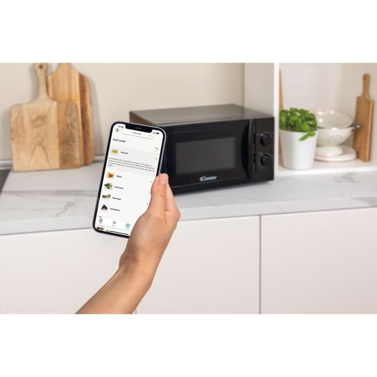 Ręka trzymająca telefon z aplikacją z przepisami, kuchenka mikrofalowa i blat.