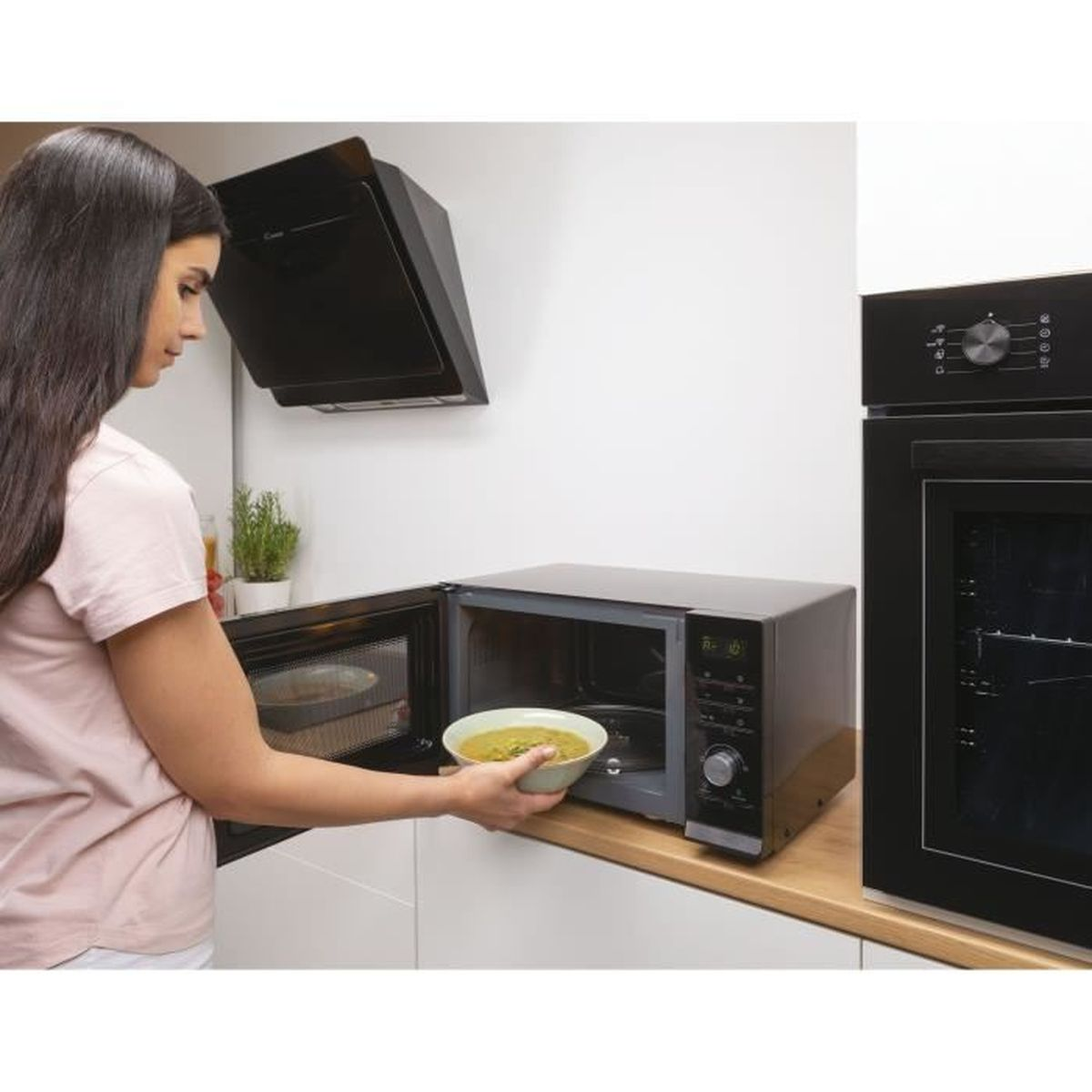 Frau legt eine Schüssel mit Essen in die Mikrowelle, im Hintergrund Ofen und Dunstabzugshaube.