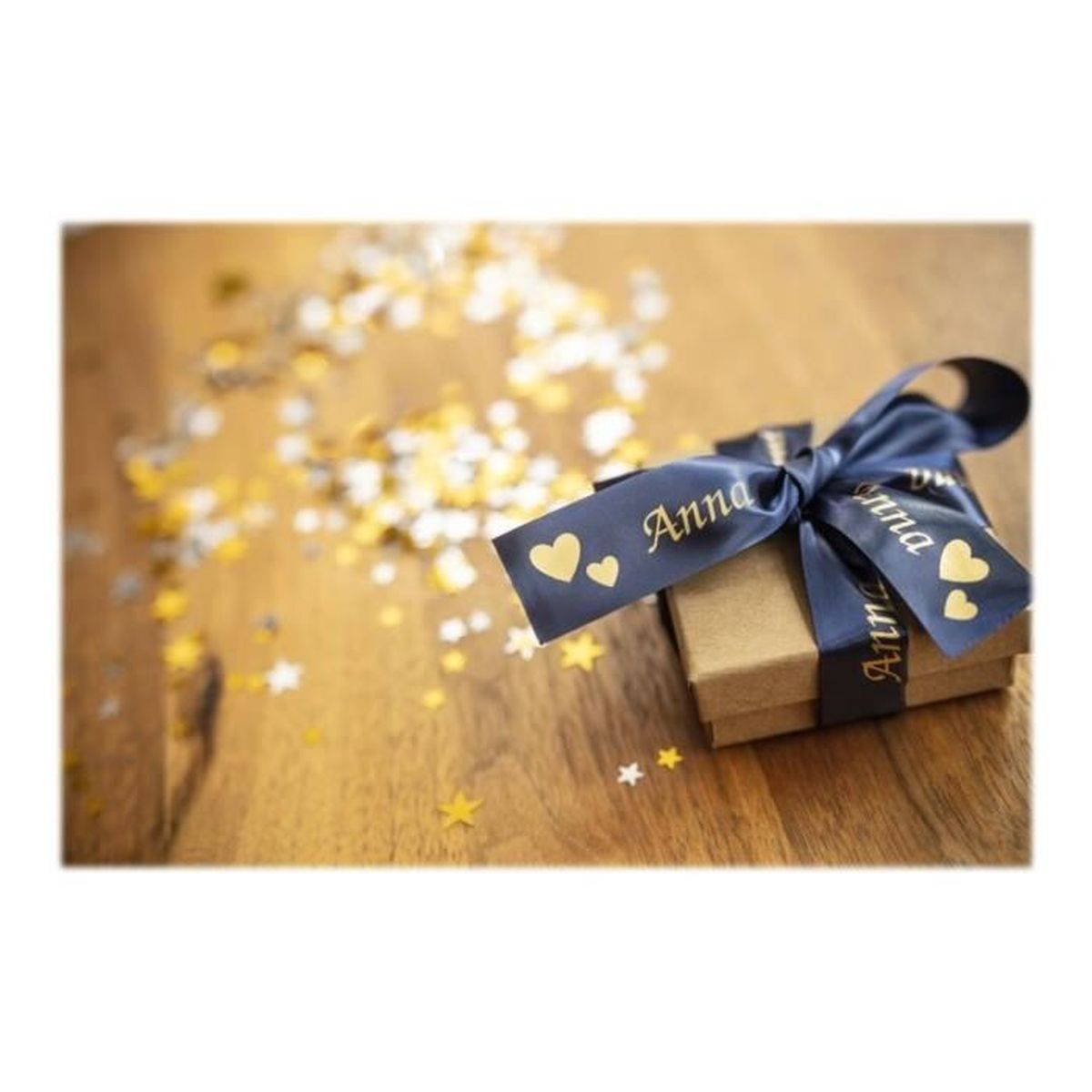Eine Geschenkbox mit einem marineblauen Band mit der Aufschrift 'Anna' und Herzen. Goldkonfetti auf dem Holztisch.
