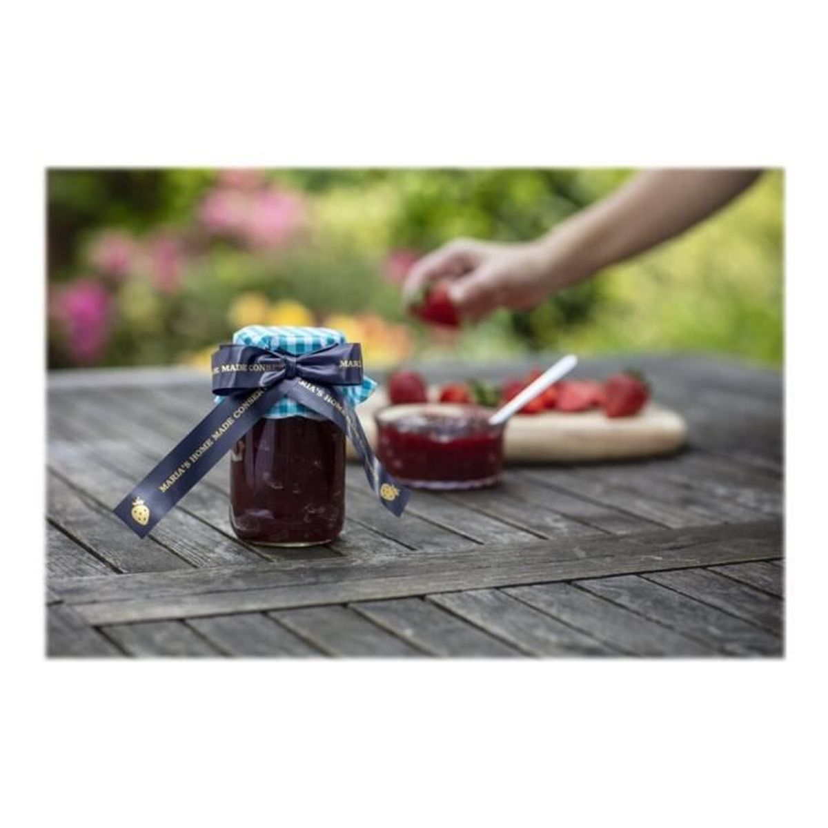 Ein Glas Marmelade mit blauem Band. Beeren und ein Löffel auf Holzbrett. Hand greift nach Beere.