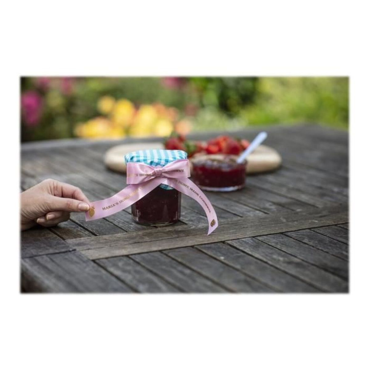 Ręka trzyma słoik dżemu, z różową wstążką wokół. Tło: słoiki i truskawki.