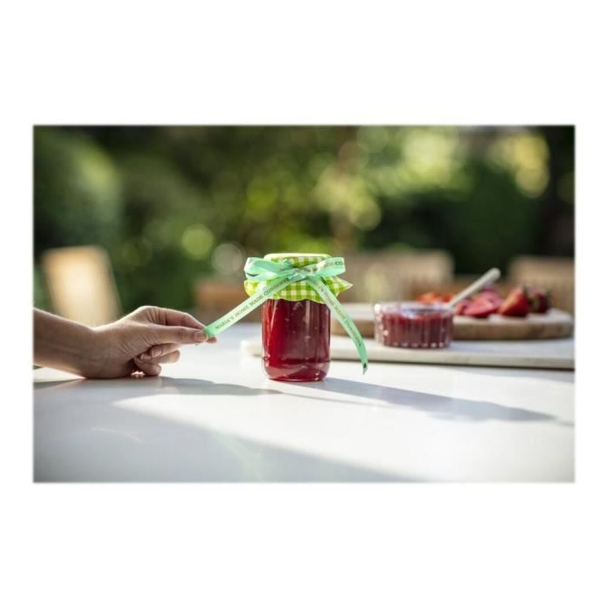 Ein Glas Marmelade mit grünem Band auf einem weißen Tisch, mit Erdbeeren und Garten im Hintergrund.