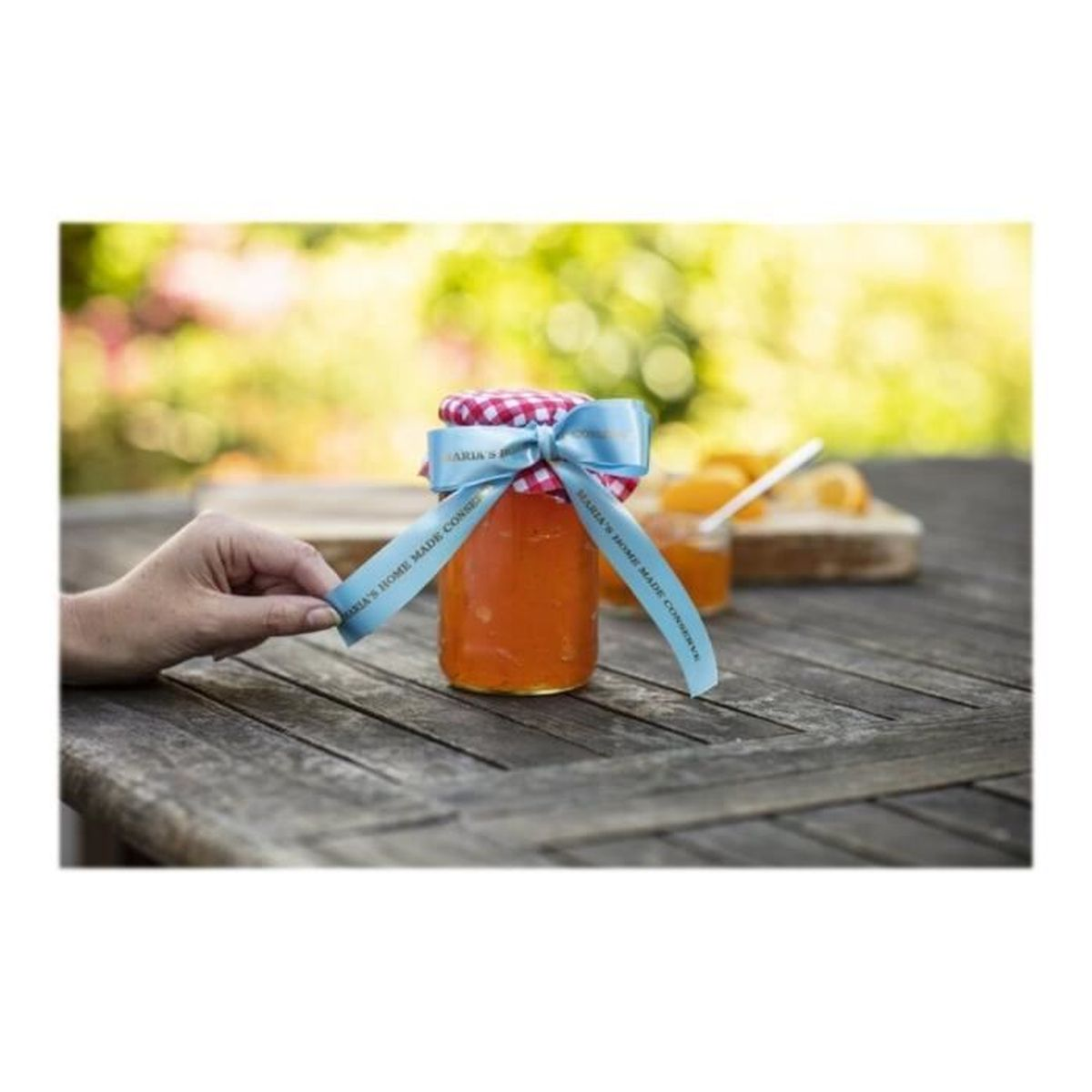 Glas mit Orangenmarmelade und blauem Band, Hand hält ein Band. Holztisch, Umgebung im Freien.