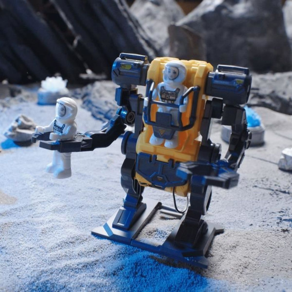 Spielzeug-Astronauten und ein Mech auf einer Mondoberfläche. Ein Astronaut wird vom Mech gehoben.