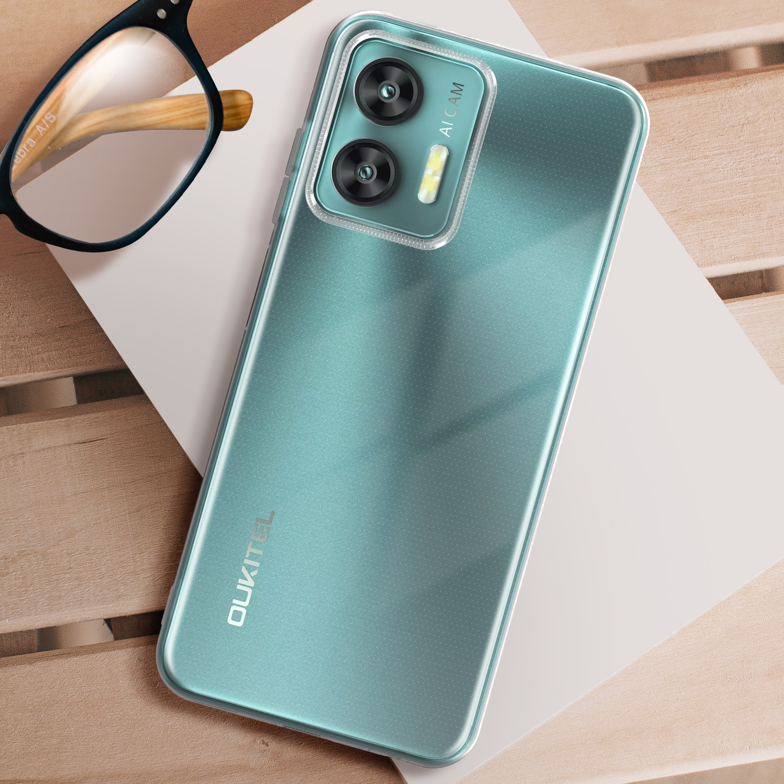 Un telefono verde acqua con doppia fotocamera, custodia trasparente e occhiali su legno.