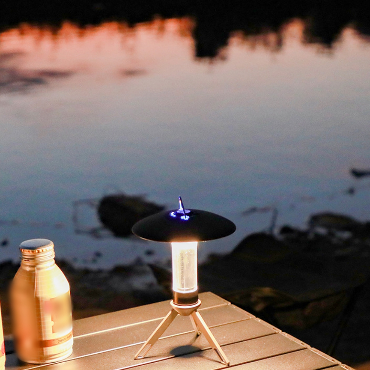 Eine Lampe auf einem Stativ, neben einer Flasche, bei Dämmerung am Wasser.