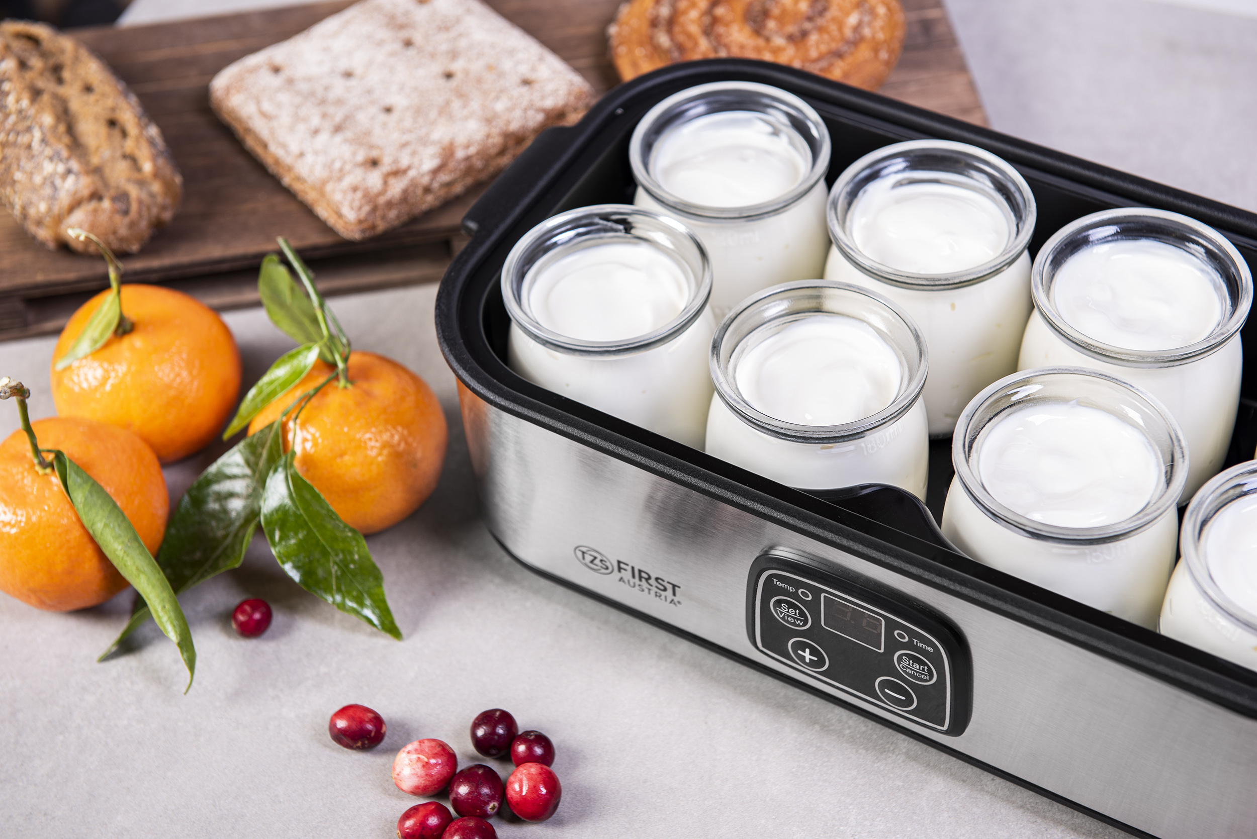 Joghurtbereiter mit gefüllten Gläsern, Orangen und Preiselbeeren. Brot und Kuchen im Hintergrund.