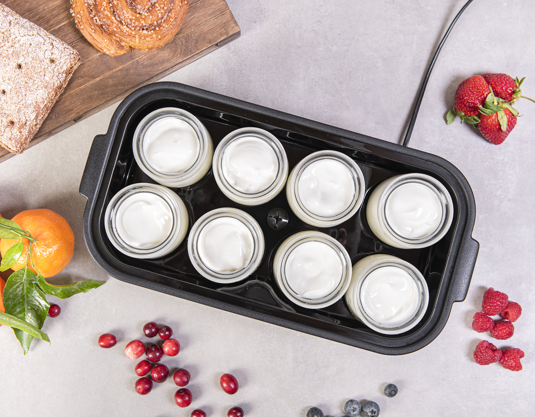 Joghurtbereiter mit acht Gläsern weißem Joghurt. Früchte und Brot liegen daneben.