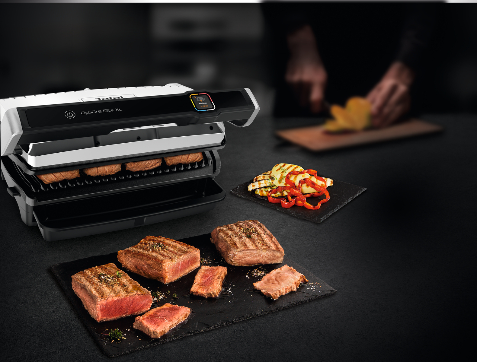 Grill mit Steaks, Fleisch und Gemüse auf dem Tisch. Eine Person ist im Hintergrund.