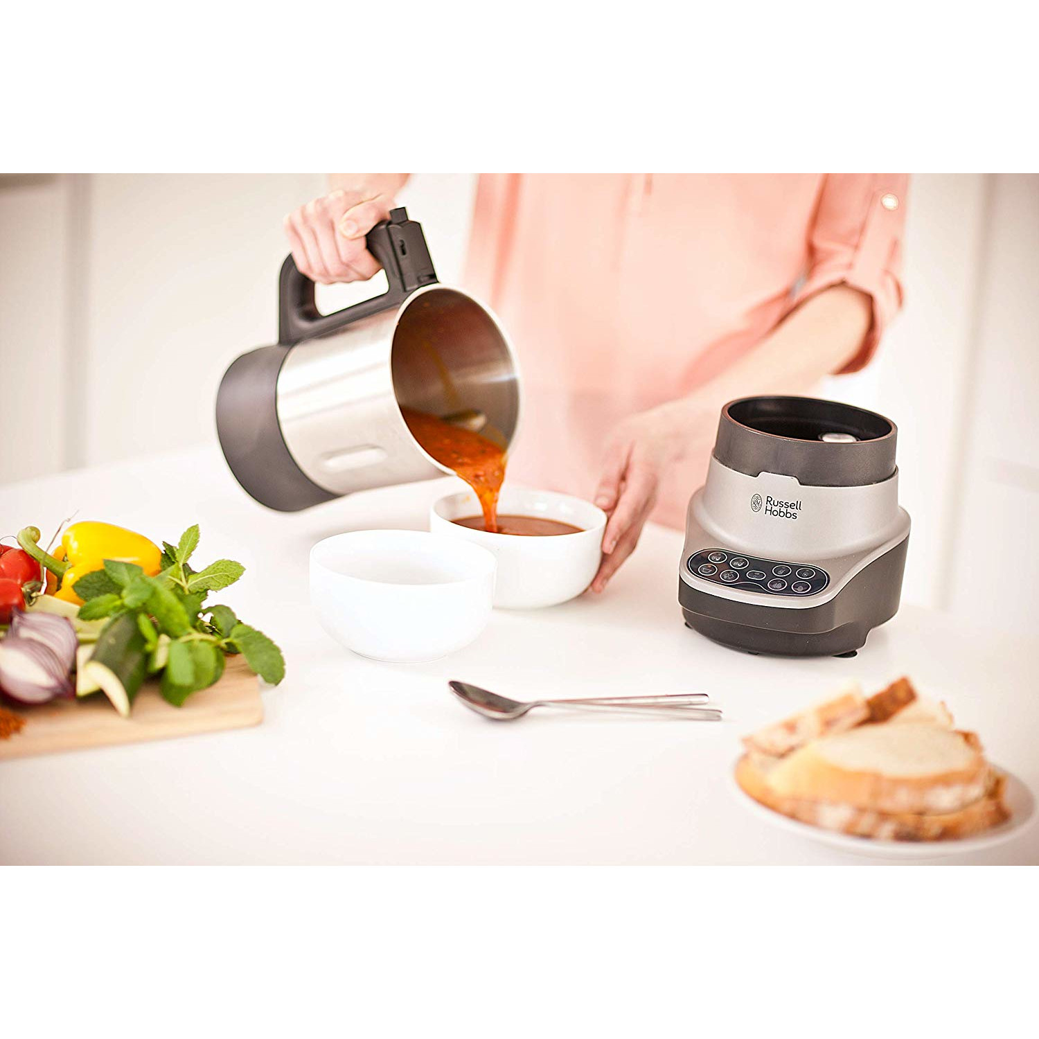 Une personne verse de la soupe d'un pichet en métal dans deux bols blancs. Un mixeur est à proximité.