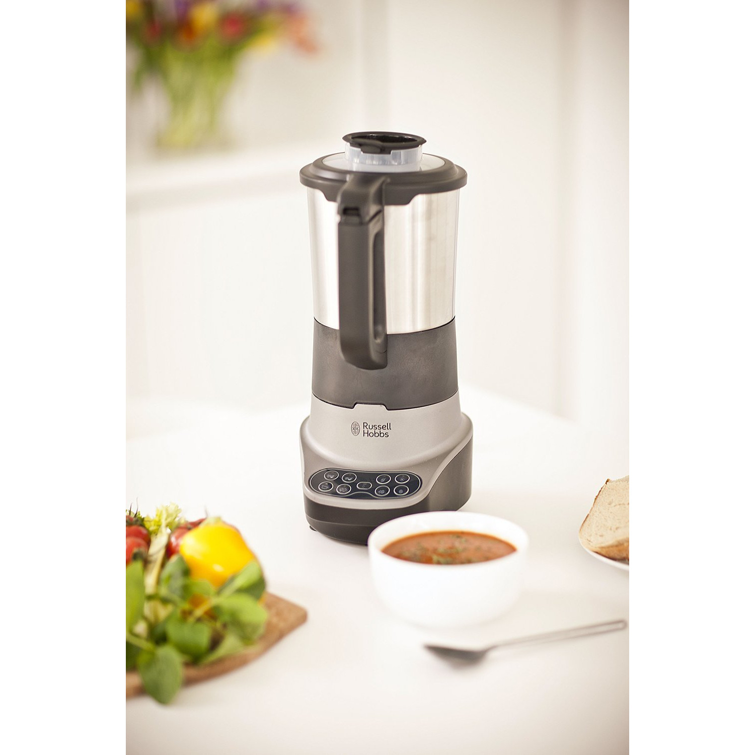 Un blender argenté et noir avec une poignée noire sur une table blanche. Un bol de soupe est à proximité.