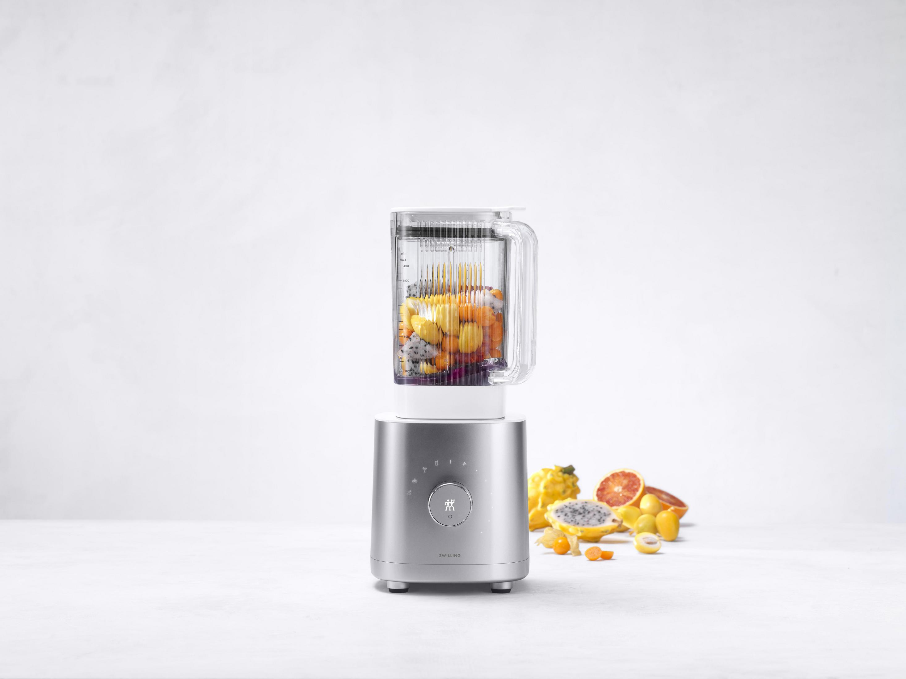 Blender z owocami w środku. Srebrna podstawa, przezroczysty pojemnik wypełniony żółtymi i pomarańczowymi owocami.