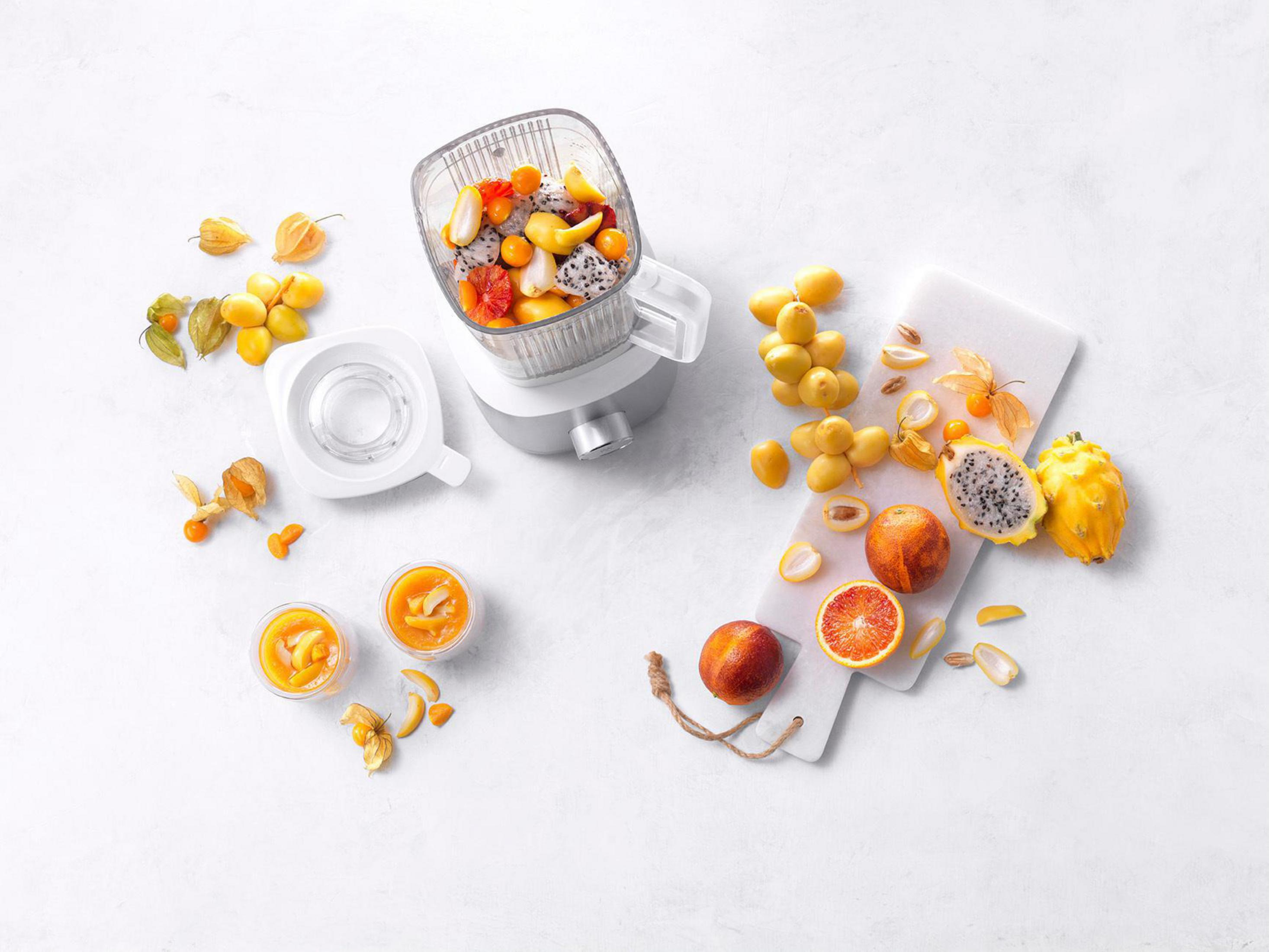 Blender z owocami, sokiem pomarańczowym w szklankach i pokrojonymi owocami na białej powierzchni.