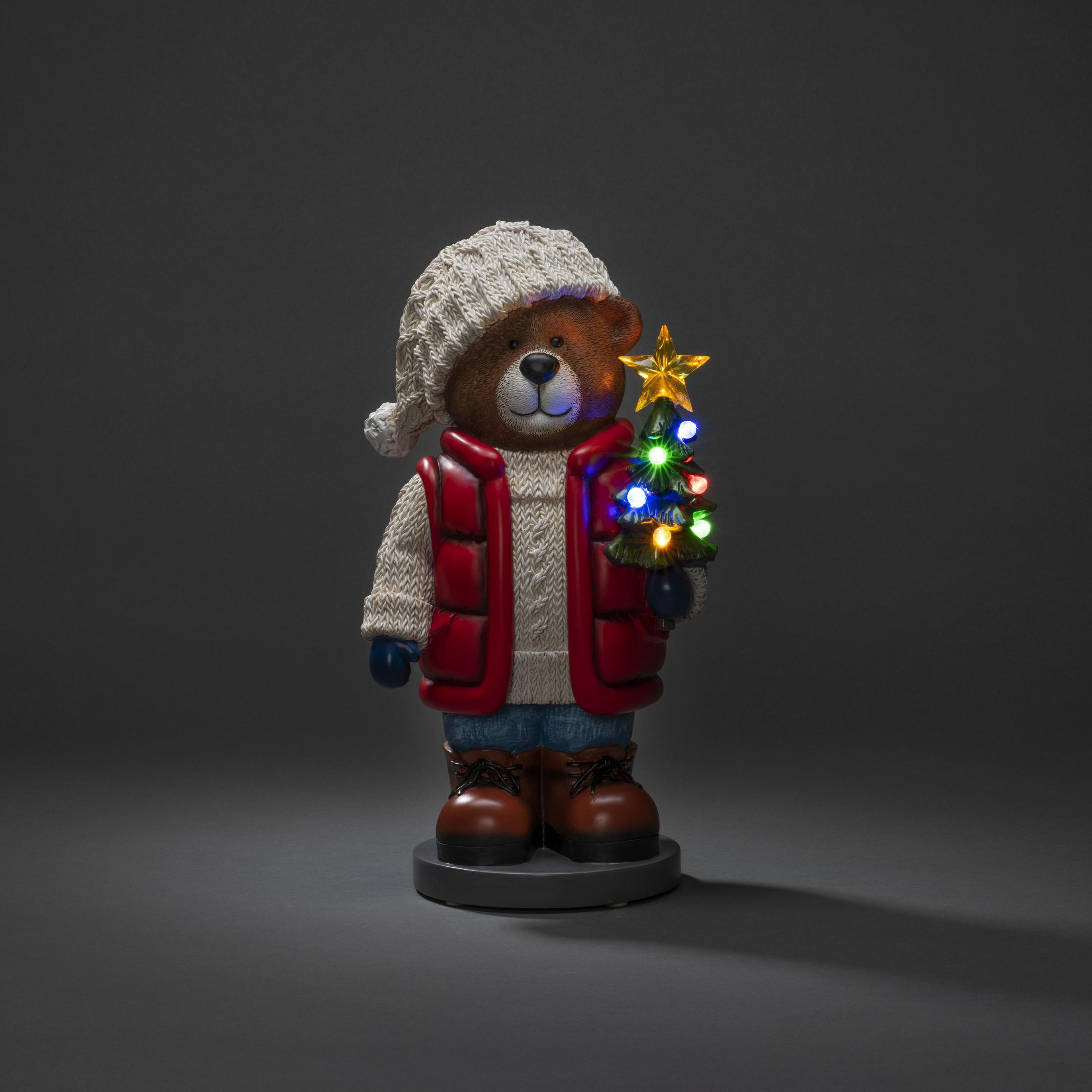 Teddybär in Winterkleidung mit beleuchtetem Weihnachtsbaum. Dunkler Hintergrund.