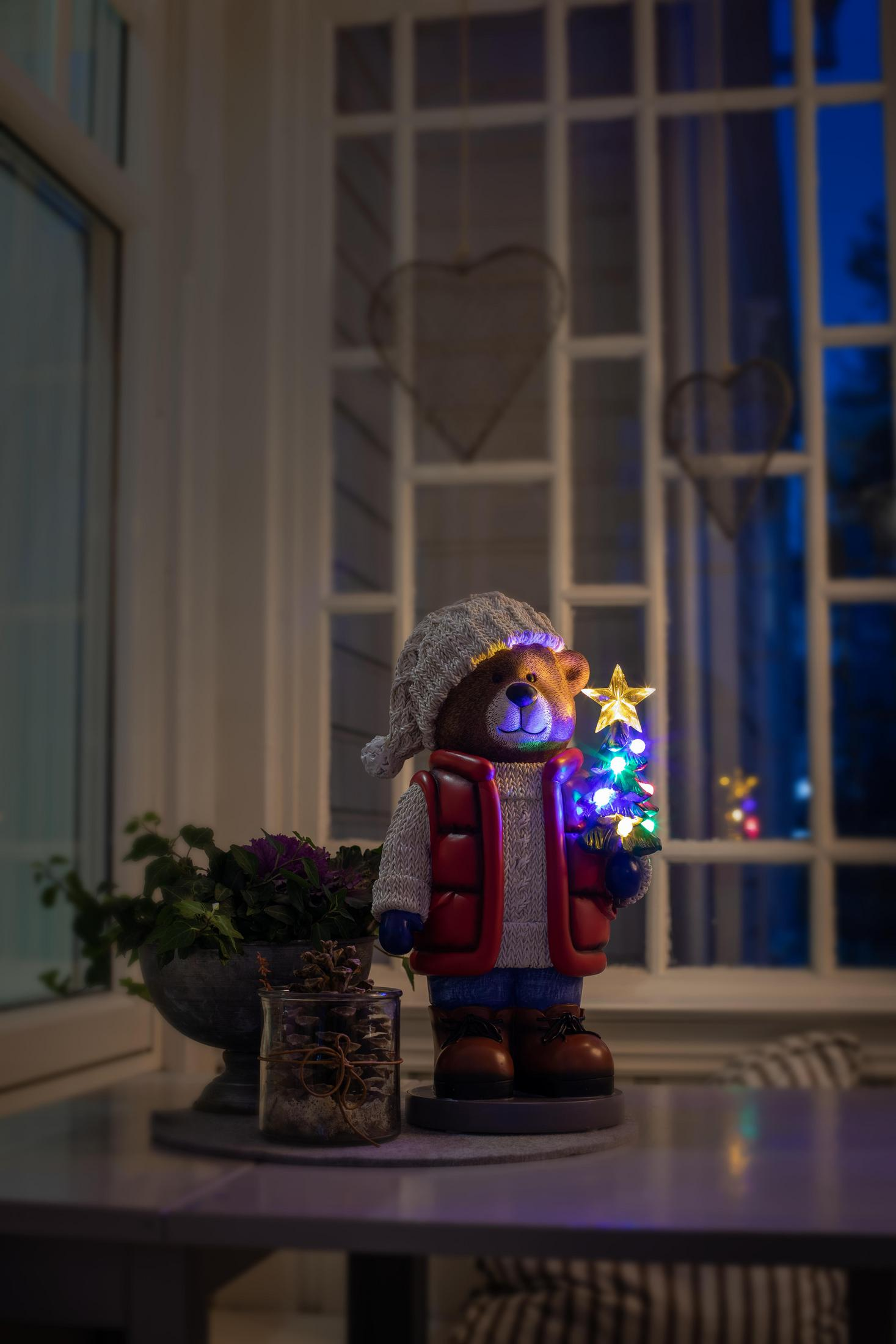 Eine Bärenfigur mit Hut und Weste, die einen Weihnachtsbaum und Stern hält, Nachtszene.