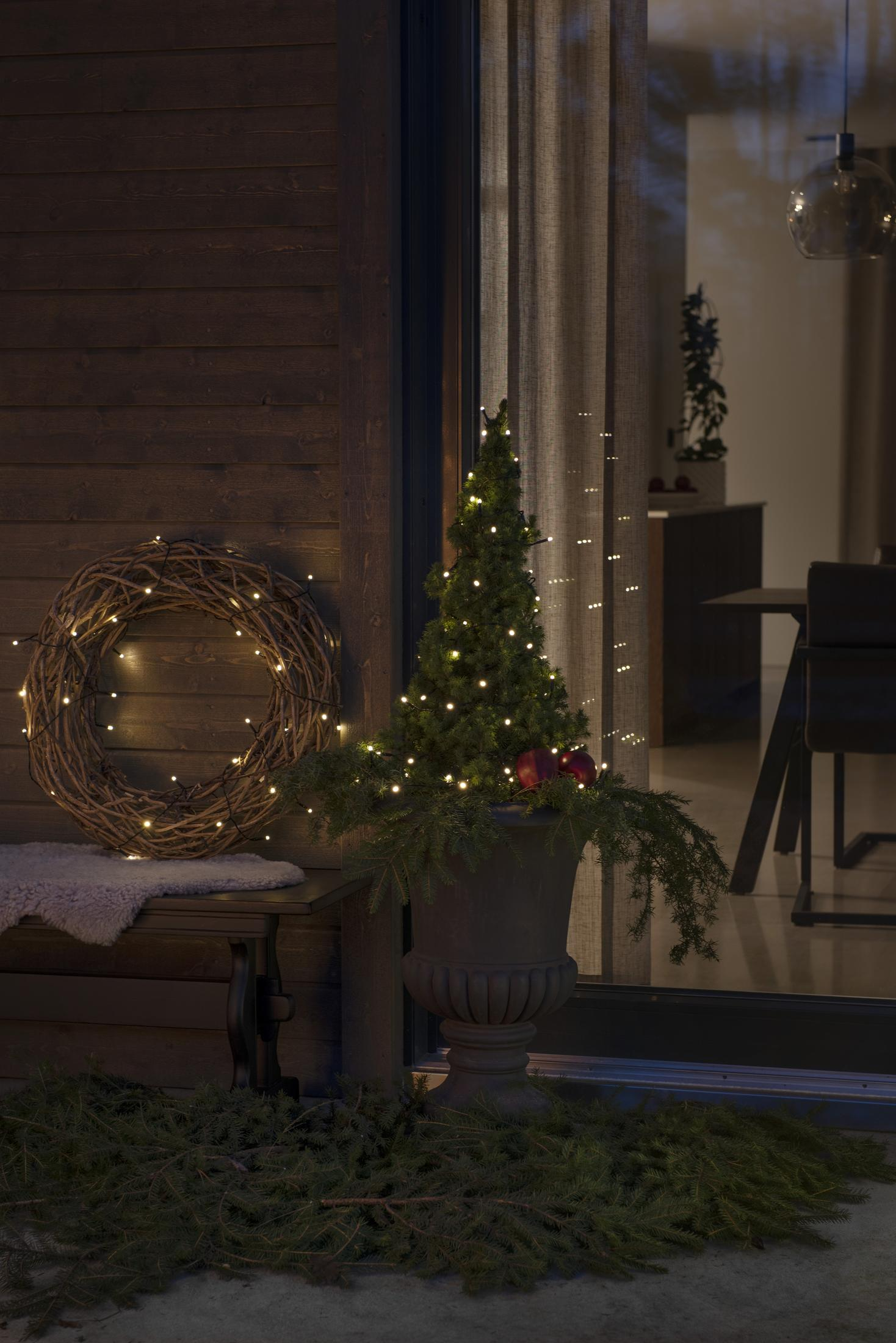 Eine Weihnachtsszene mit einem Kranz, einem Weihnachtsbaum in einem Topf und Lichtern auf einer Veranda mit einer Holzwand.
