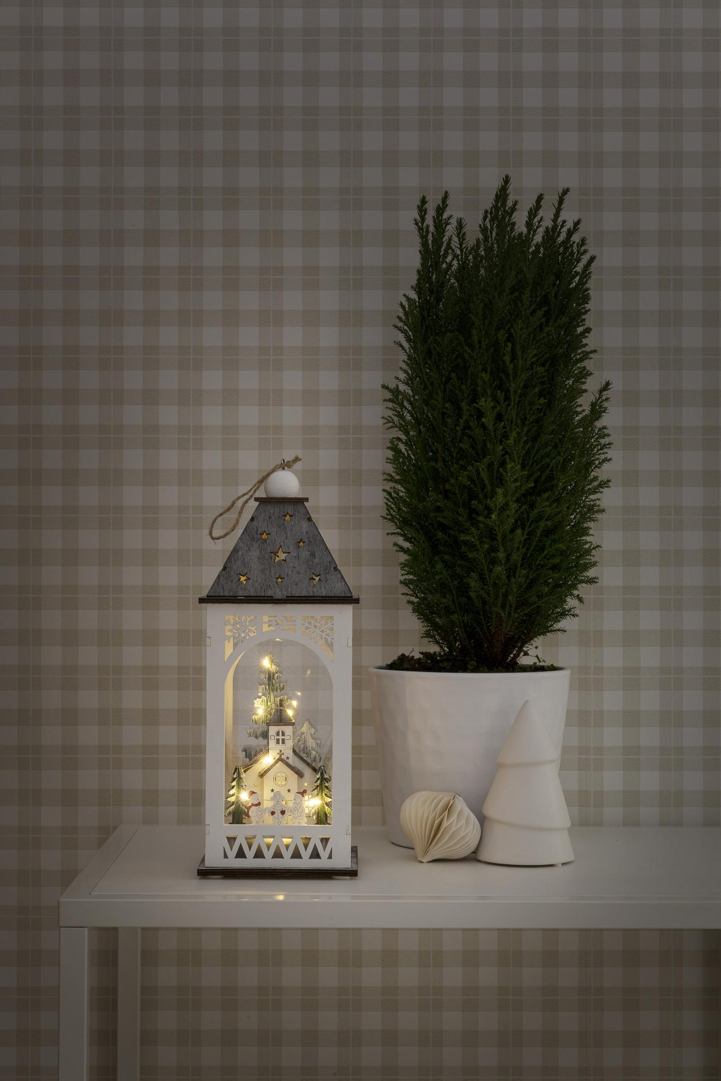 Weihnachtslaterne mit Lichtern, kleinem Baum und einer Pflanze auf einem Tisch vor einer karierten Wand.