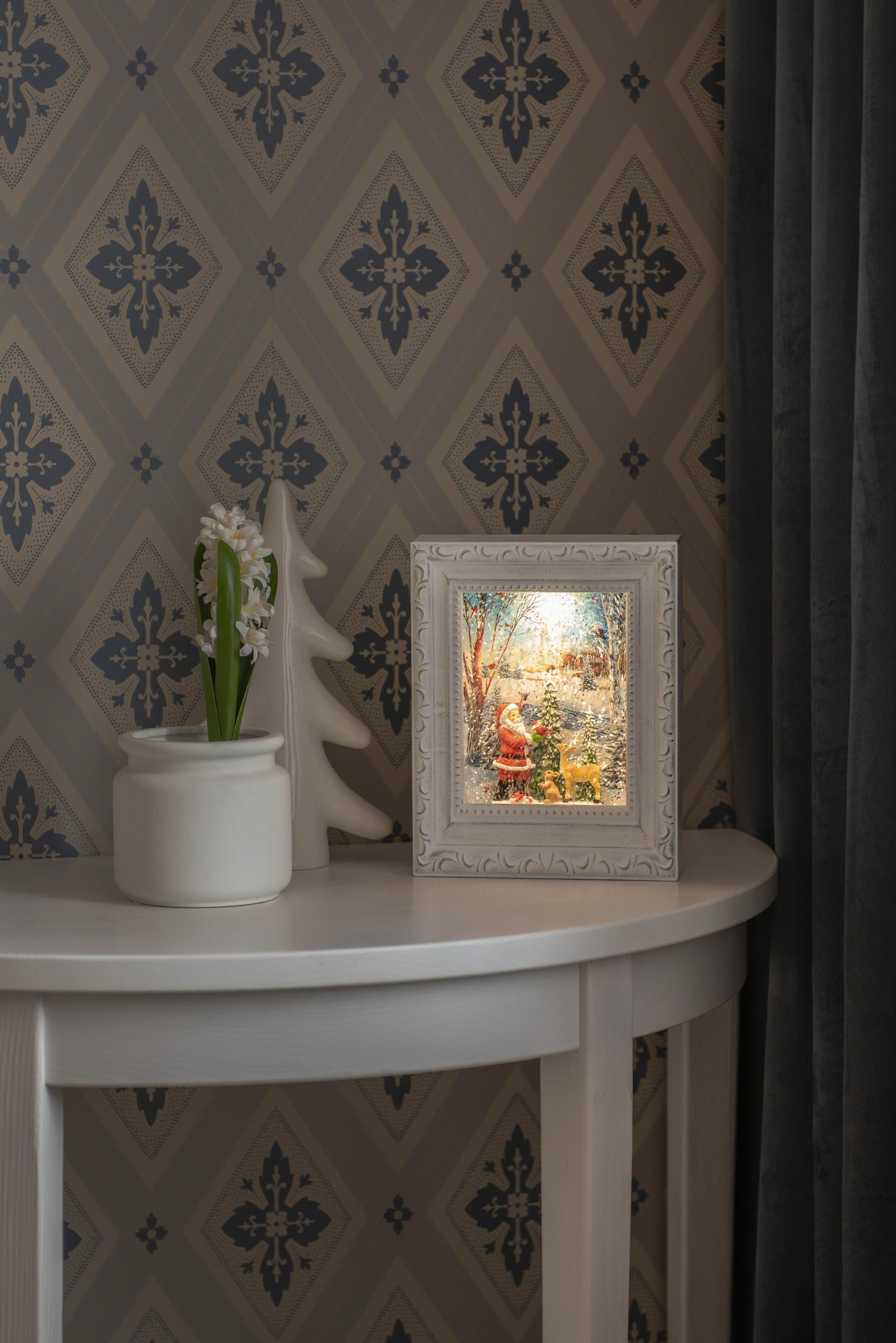 Kleine Weihnachtsszene in einem Rahmen auf einem Tisch. Ein weißer Baum und eine Vase mit Blumen sind in der Nähe.