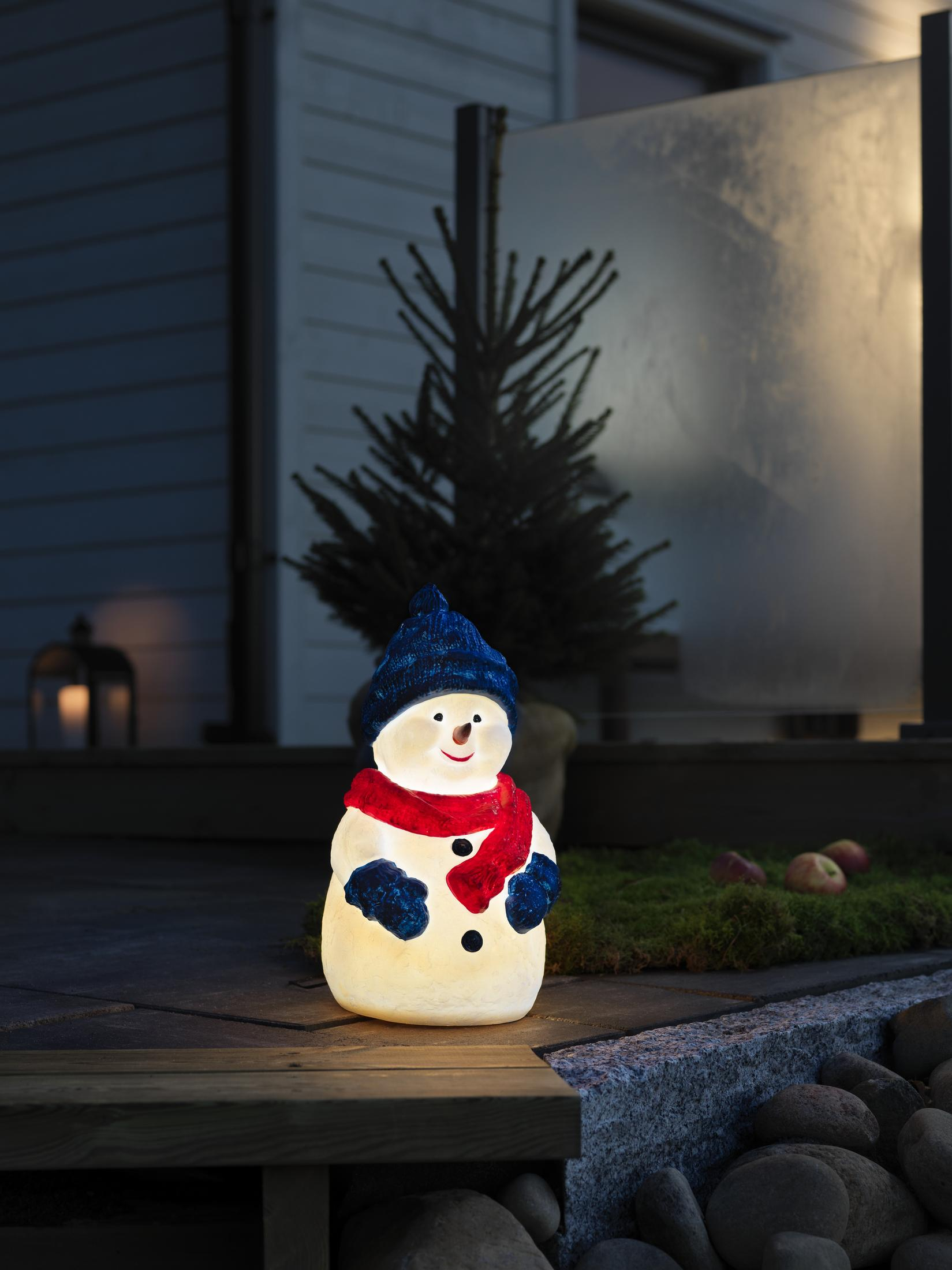 Schneemann-Statue mit blauem Hut und rotem Schal auf beleuchteter Veranda.