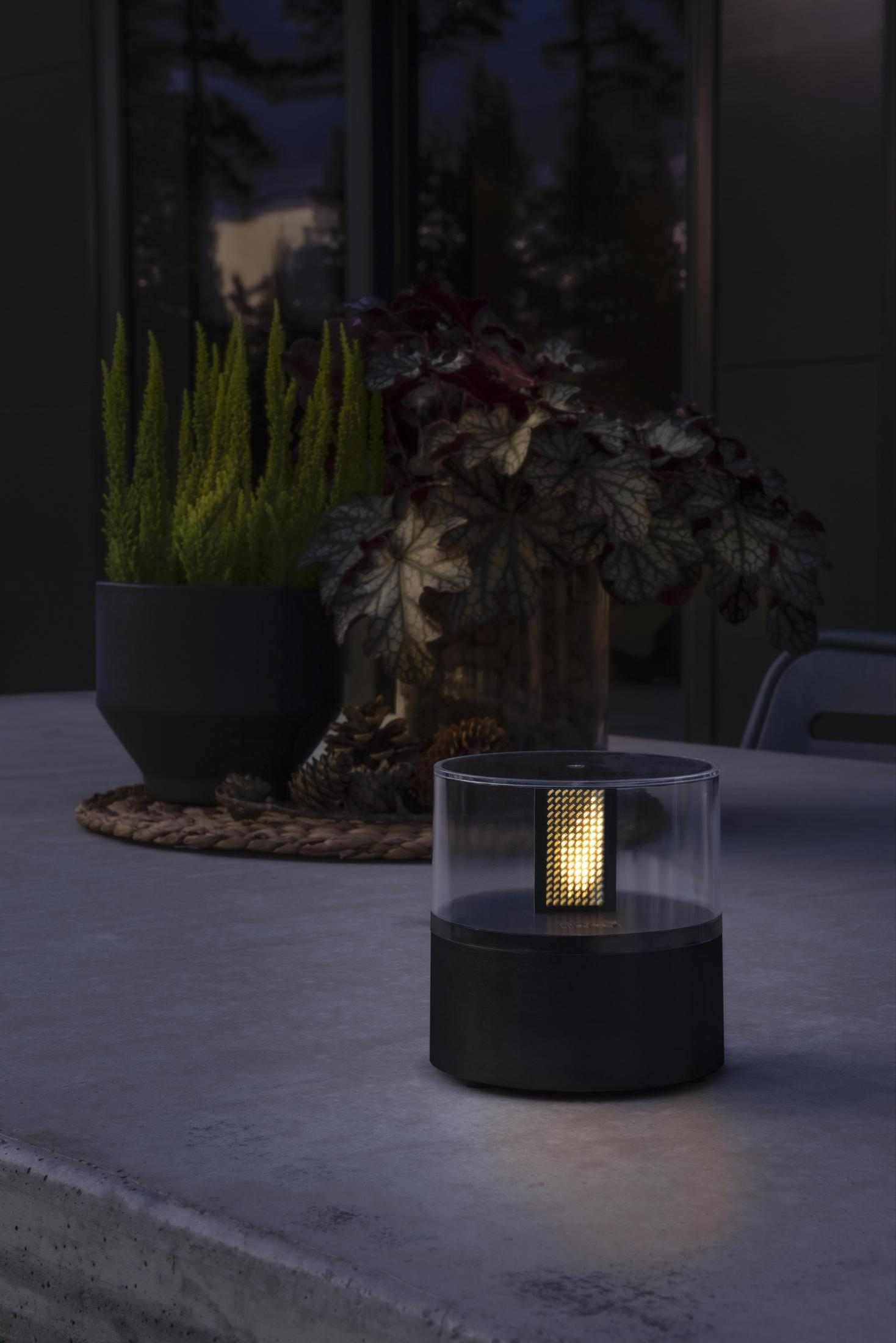Eine beleuchtete Außenlampe mit schwarzem Sockel. Pflanzen und ein Tisch im Hintergrund.