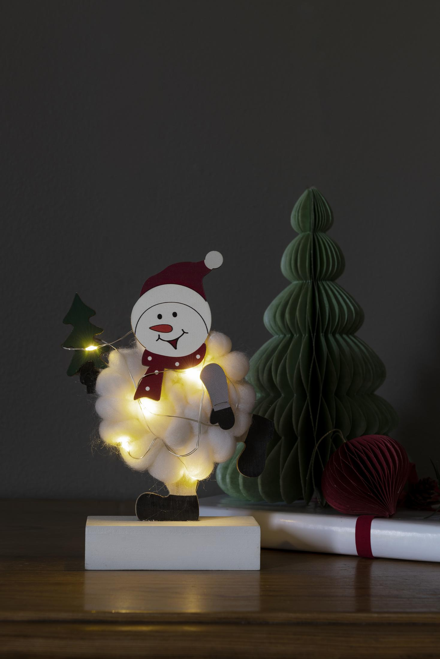 Eine Schneemann-Dekoration mit Lichtern und einem Baum. Auf Holzsockel, grauer Hintergrund.
