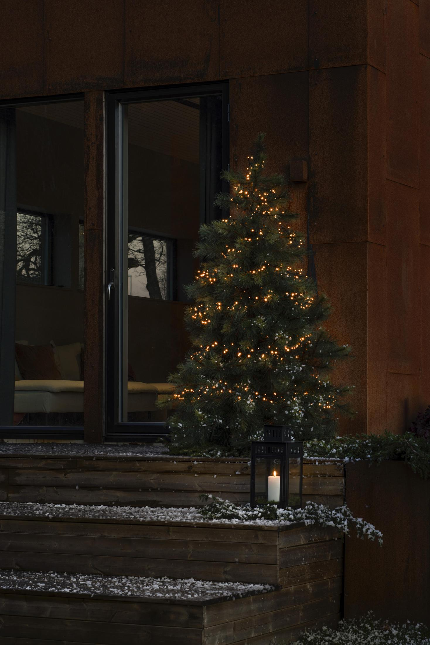 Weihnachtsszene im Freien mit einem beleuchteten Baum. Eine Kerzenlaterne auf den Holzstufen.