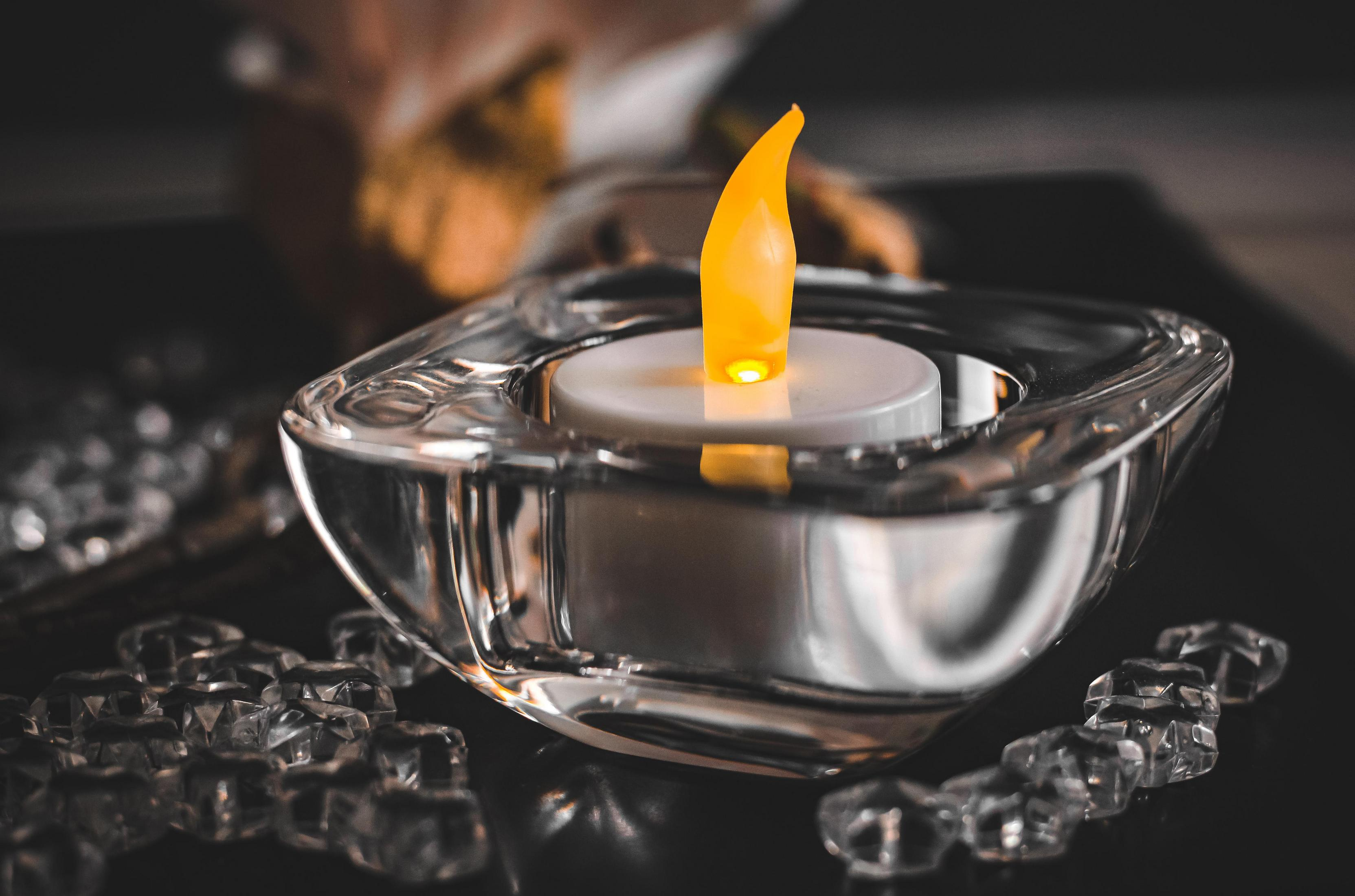 Eine brennende Kerze mit gelber Flamme in einem Glasbehälter, umgeben von Kristallen auf schwarzem Untergrund.