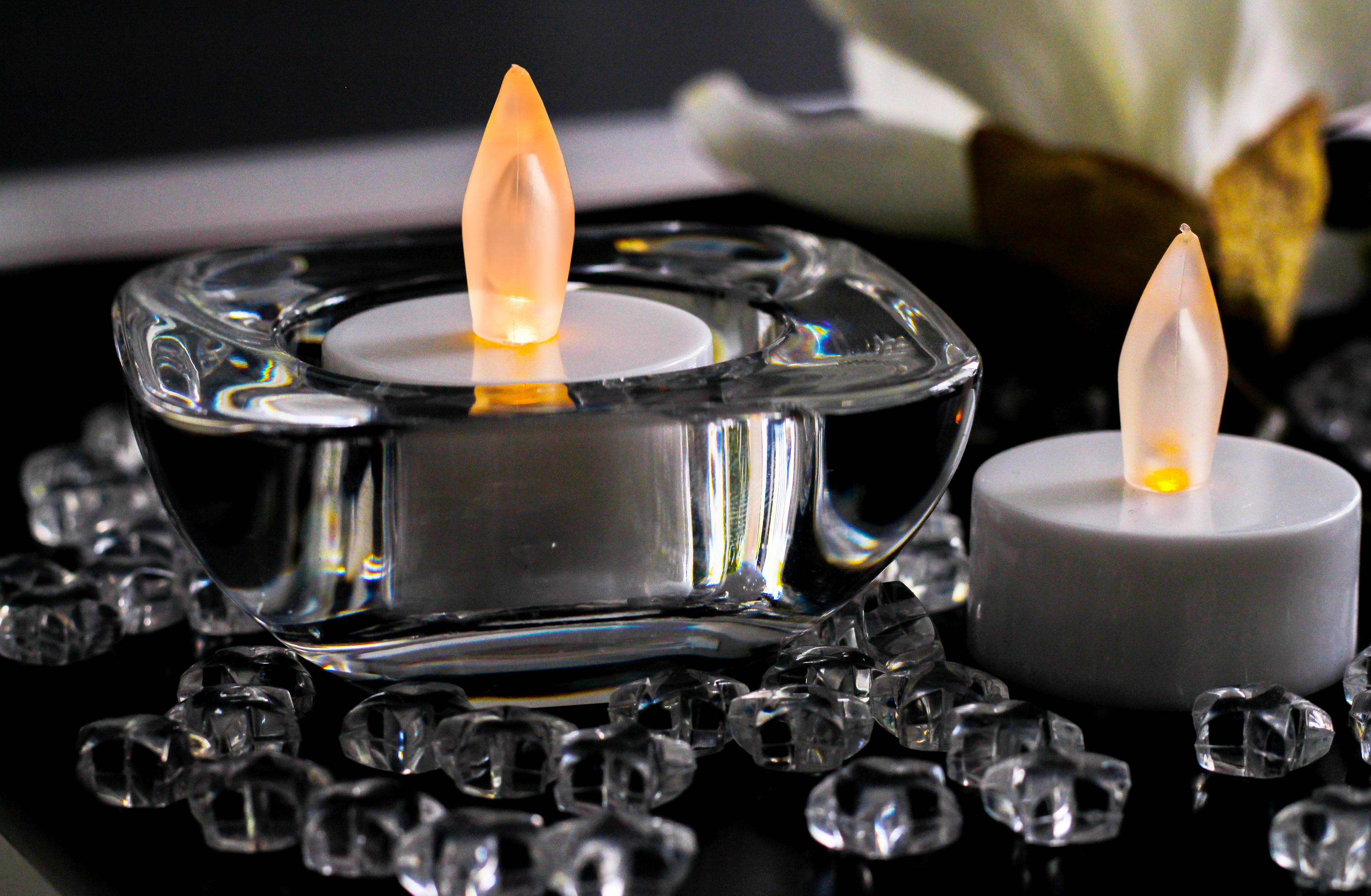 Zwei Kerzen mit flammenförmigen Lichtern, in einem klaren Glasbehälter und auf weißer Oberfläche.