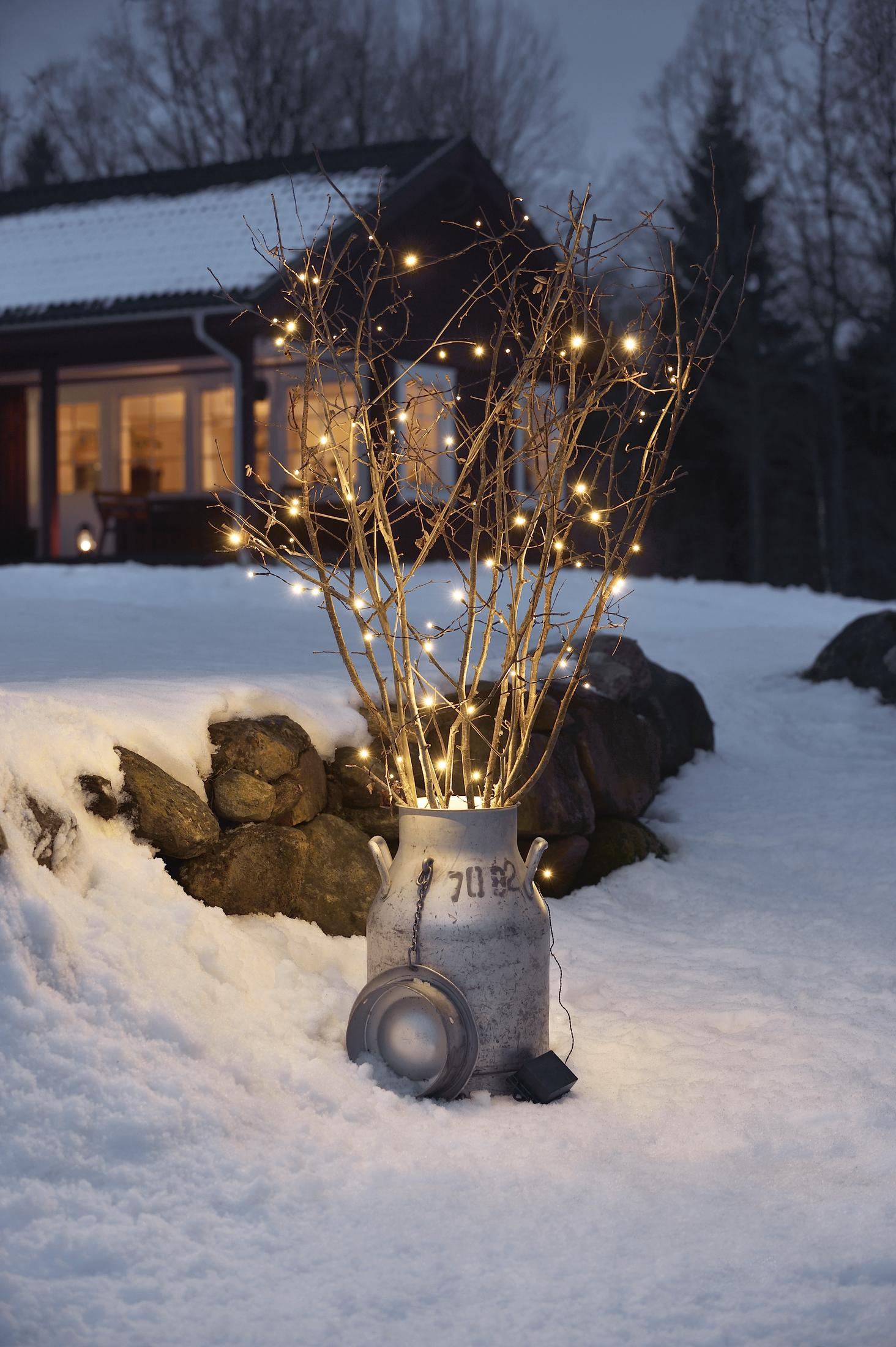 Beleuchtete Zweige in einem Metallbehälter in einem verschneiten Garten, mit einem Haus im Hintergrund bei Dämmerung.