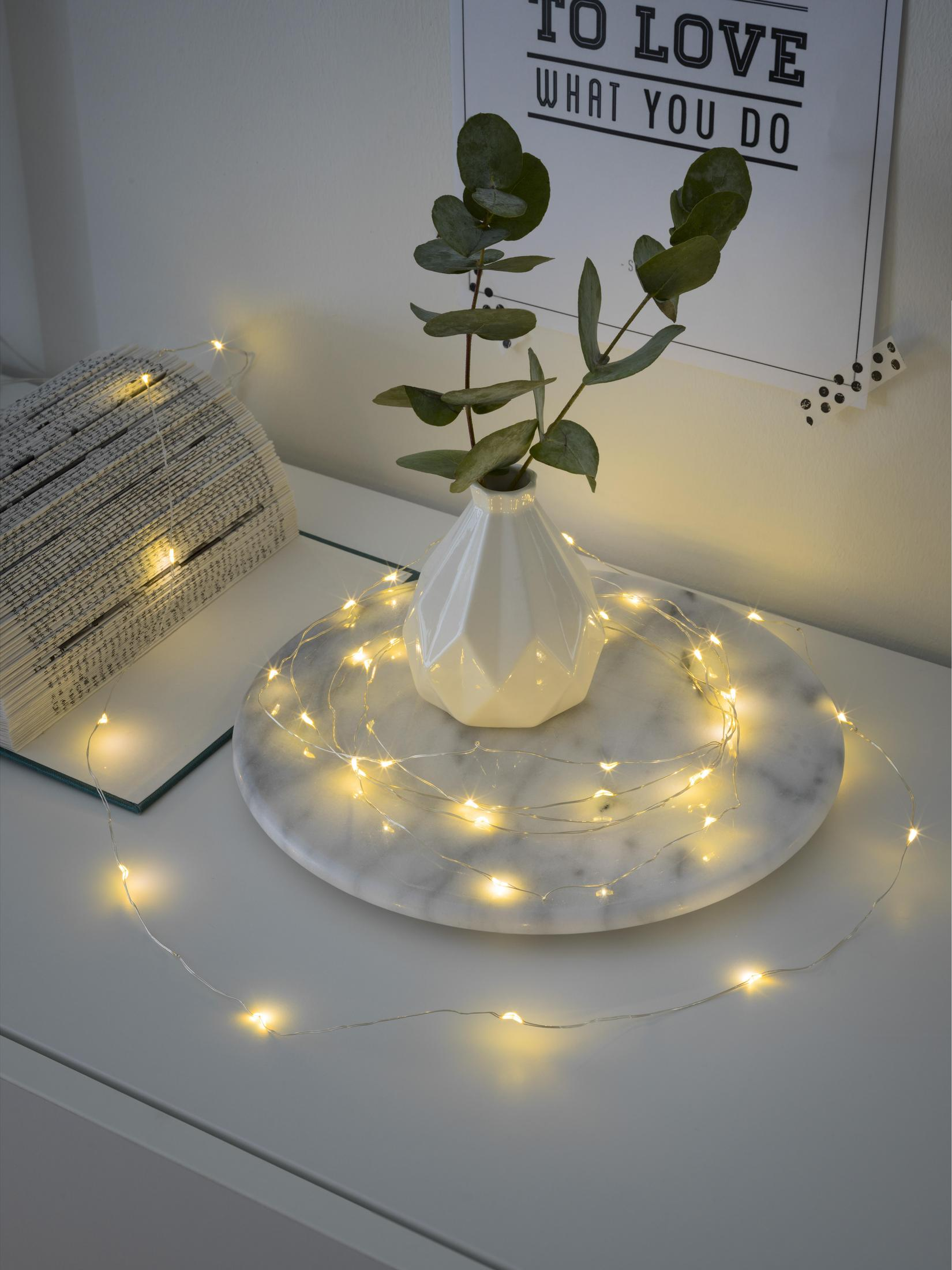 Eine weiße Vase mit grünen Blättern und Lichterketten auf einem Marmortablett. Ein Buch liegt daneben.