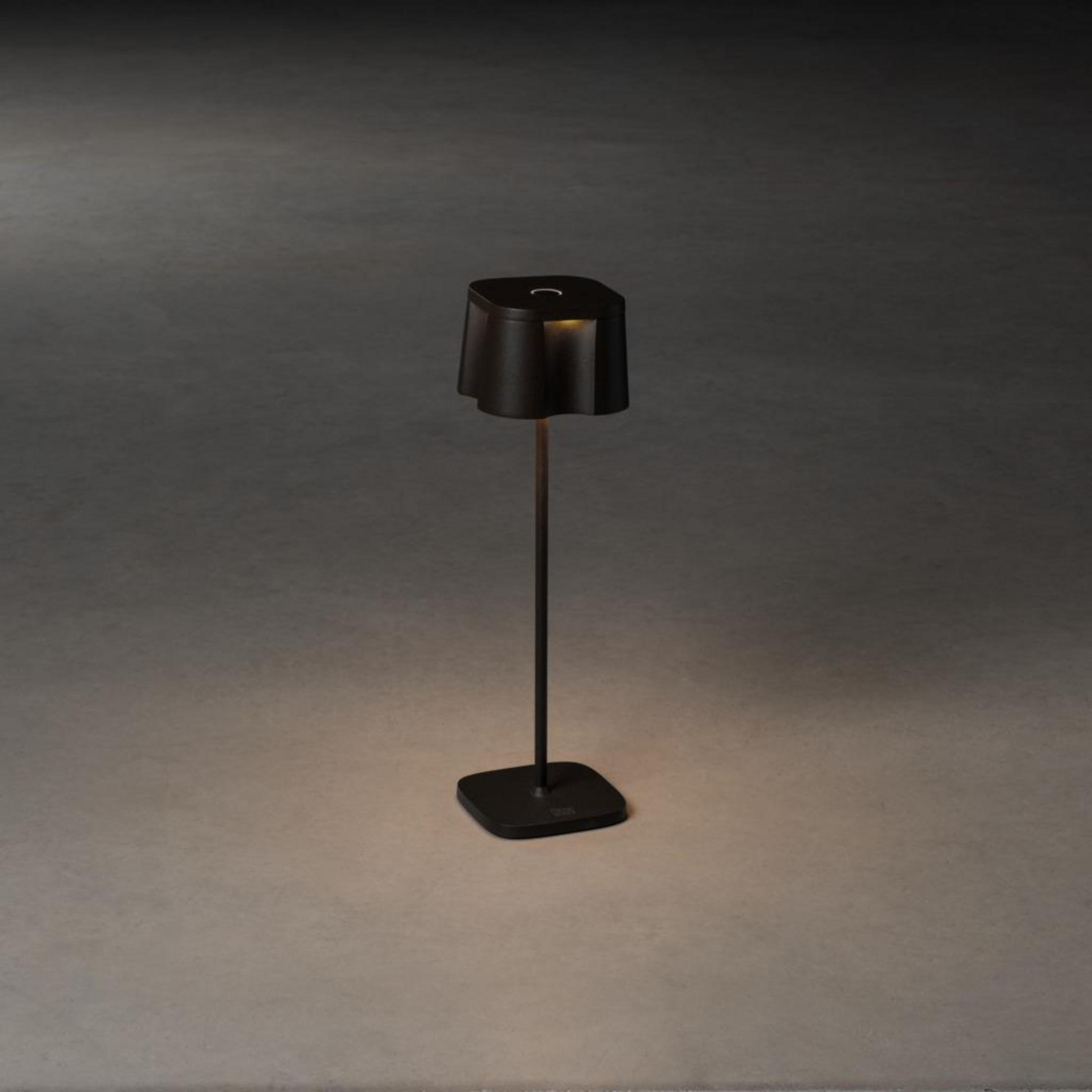 Eine schwarze Lampe mit hohem Ständer und quadratischem Sockel. Die Lampe ist an.