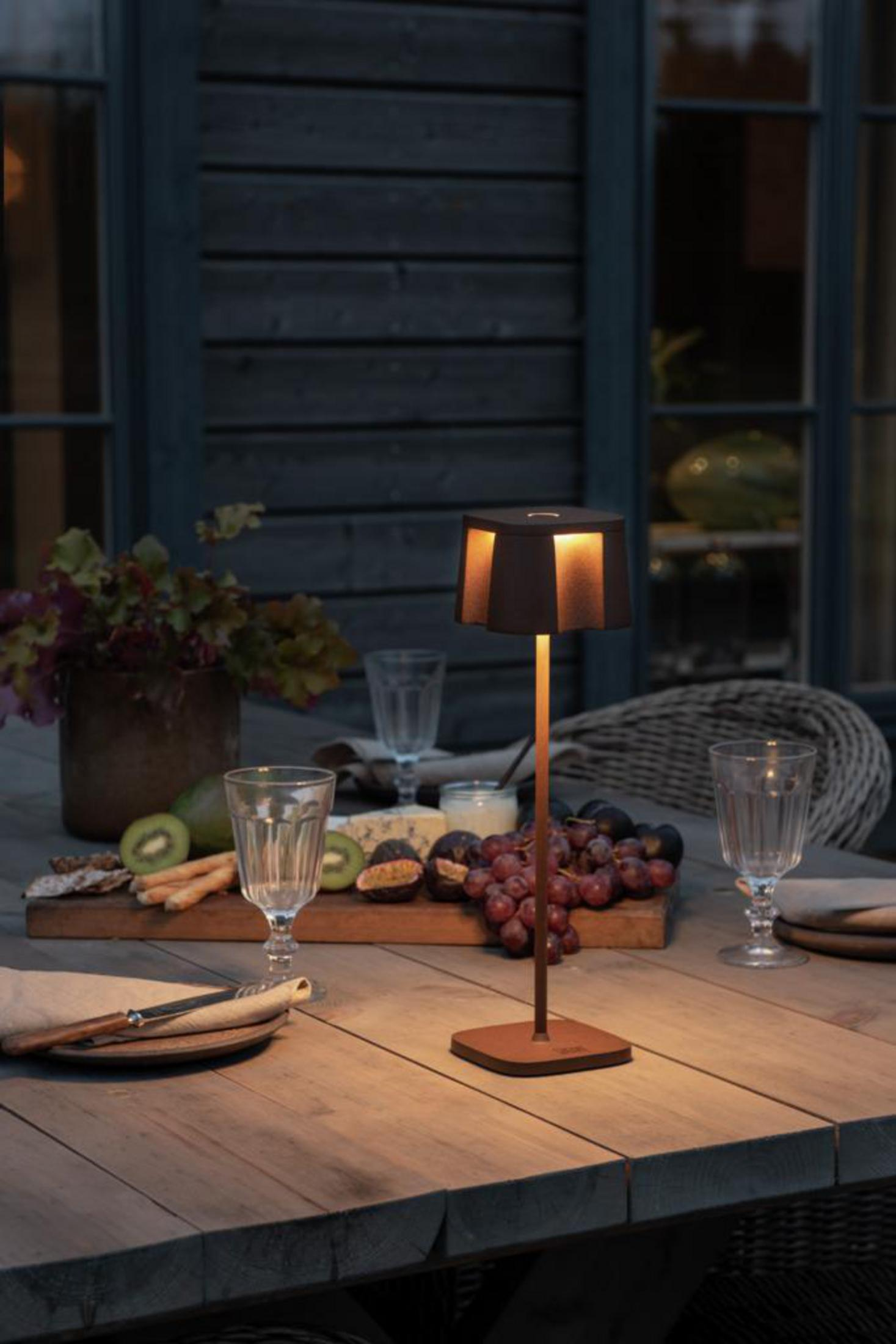 Eine Tischlampe mit schwarzem Schirm und warmem Licht, auf einem Holztisch mit Essen.